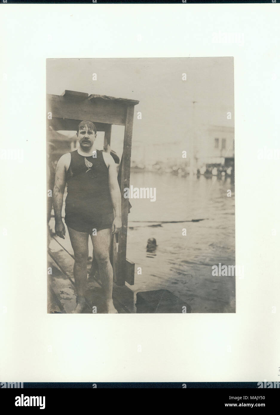 Wet man in a swimsuit standing on a wooden dock beside a small lake with a man swimming. Title: W.E. Dickey of the New York Athletic Club, as the Plunge for Distance champion at the 1904 Olympics.  . 1904. Stock Photo