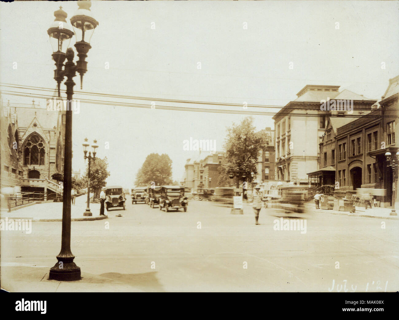 Looking west on Lindell Boulevard from Grand Avenue. Photo taken July 1, 1921. Automobiles and a traffic officer on Lindell Boulevard west of Grand Avenue. Elegant private homes lined the north side of the 3600 block of Lindell. Saint Francis Xavier Church is pictured at left. The Berlitz School of Languages is pictured at right, at the northwest corner of the intersection. The school's building was razed in 1921 to permit the construction of the fashionable Melbourne Hotel. Covenant Blu, Midtown, Saint Louis University Title: Looking west on Lindell Boulevard from Grand Avenue.  . 1 July 1921 Stock Photo