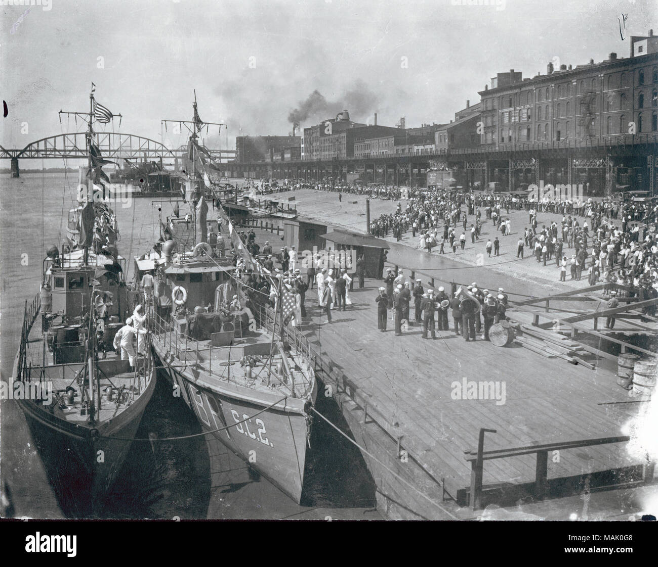 Horizontal, black and white photograph of a crowd greeting the United States Navy anti-submarine flotilla. Two flotillas are docked against a wooden deck with soldiers in both white and dark uniforms. The McKinley Bridge is present in the background of the photograph and railroad tracks are seen at the beginning of the city buildings. Title: Crowd on riverfront greeting U.S. Navy anti-submarine flotilla for demonstration visit. 13 June 1919.  . 1919. Stock Photo