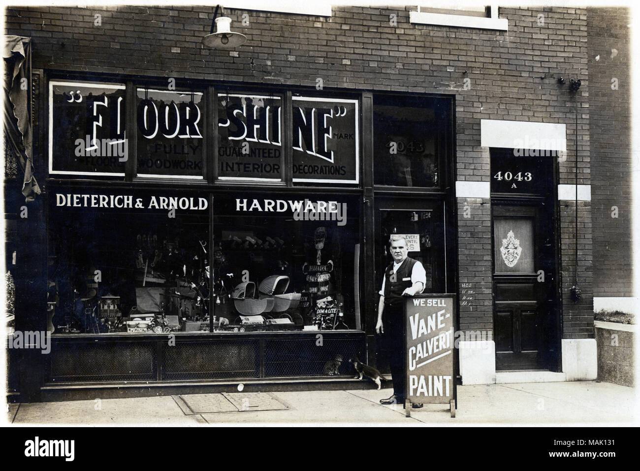 Photograph of the first story of a brick building. A man is standing in front of the store with his right arm on a sign. Title: Dietrich and Arnold Hardware Store, 4043 Lexington Avenue.  . December 1921. Stock Photo