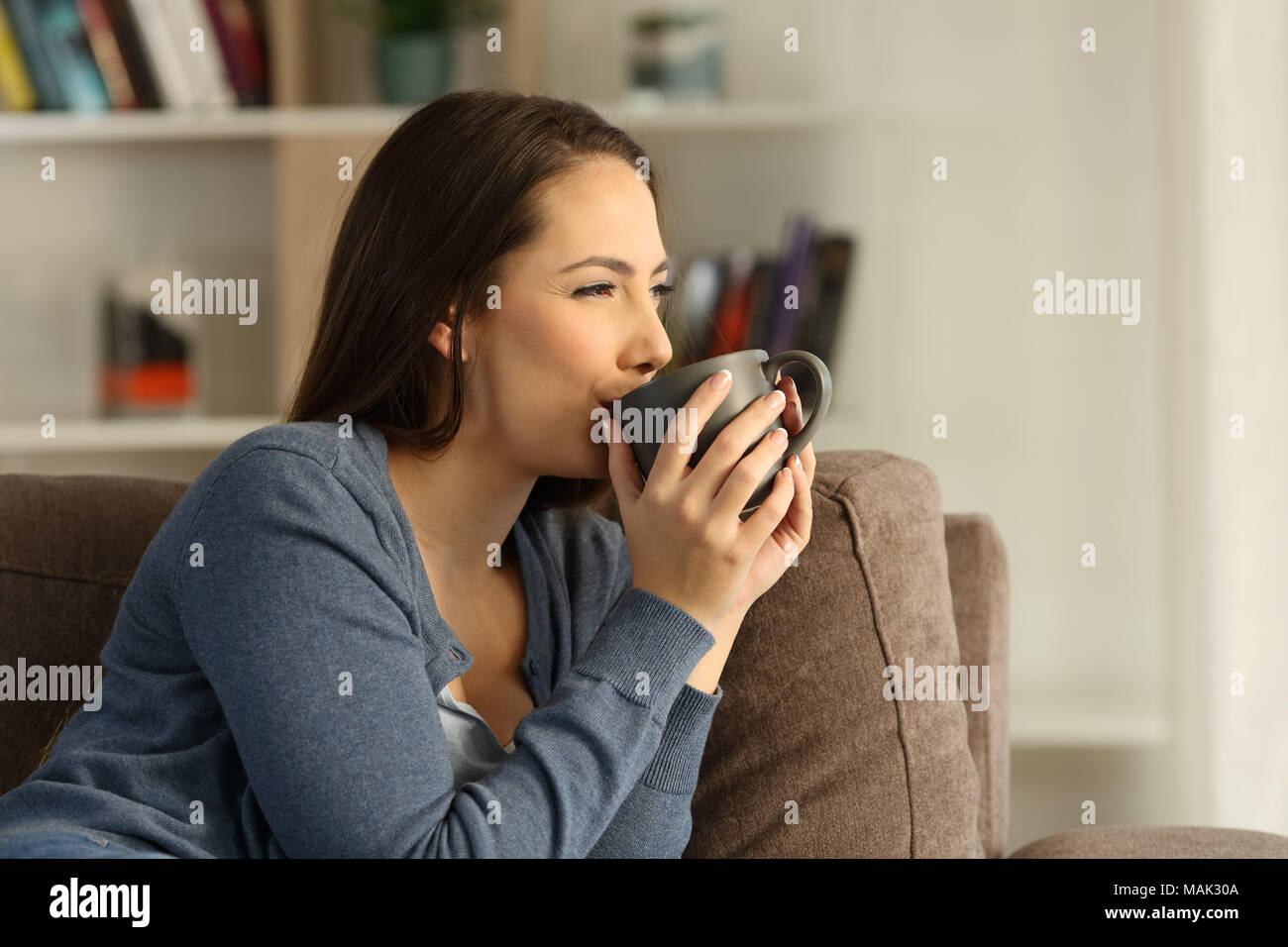 Relaxed happy woman drinking coffee and looking away sitting on a couch in the living room at home Stock Photo