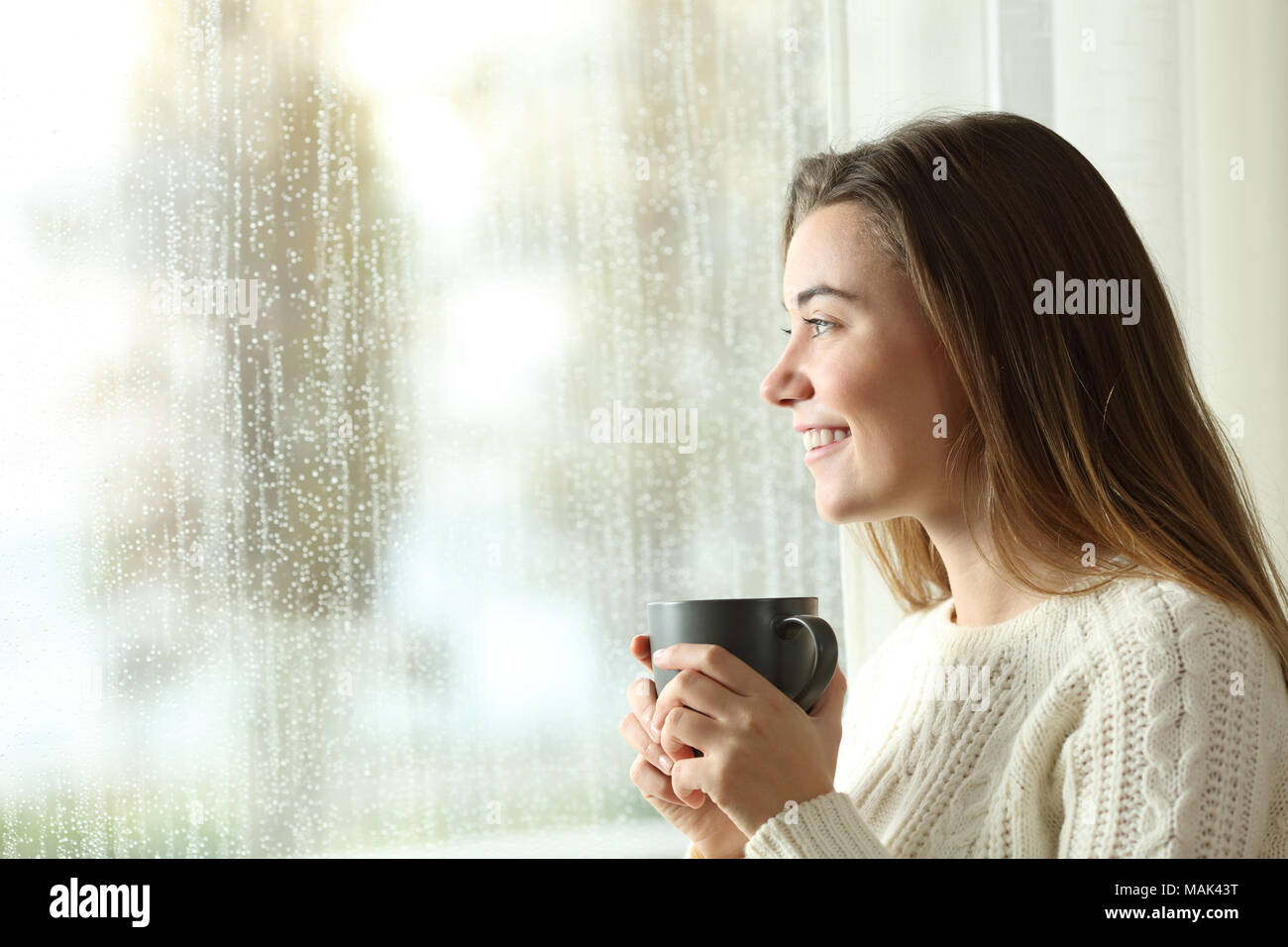 Happy teen holding a mug looking through a window at home in a rainy day Stock Photo
