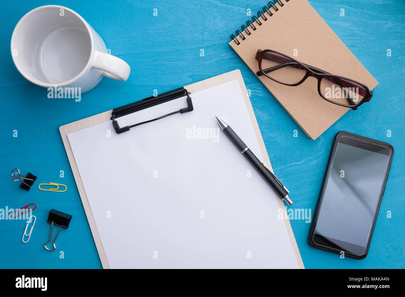 A4 size clipboard, smartphone, small notebook and coffee cup on blue wood table in workplace. background with blank area for text or message. Stock Photo