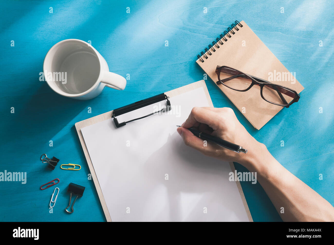 Young Asian male hand writing on white paper on a4 size clipboard on blue wood table in workplace. freelance worker lifestyle concept Stock Photo