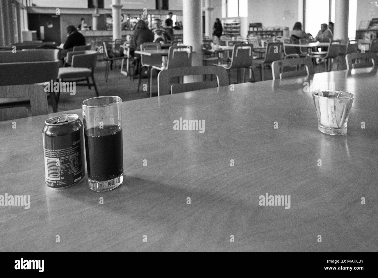 A can of cola and glass in with a cafe out of focus in the background, grainy black and white Stock Photo