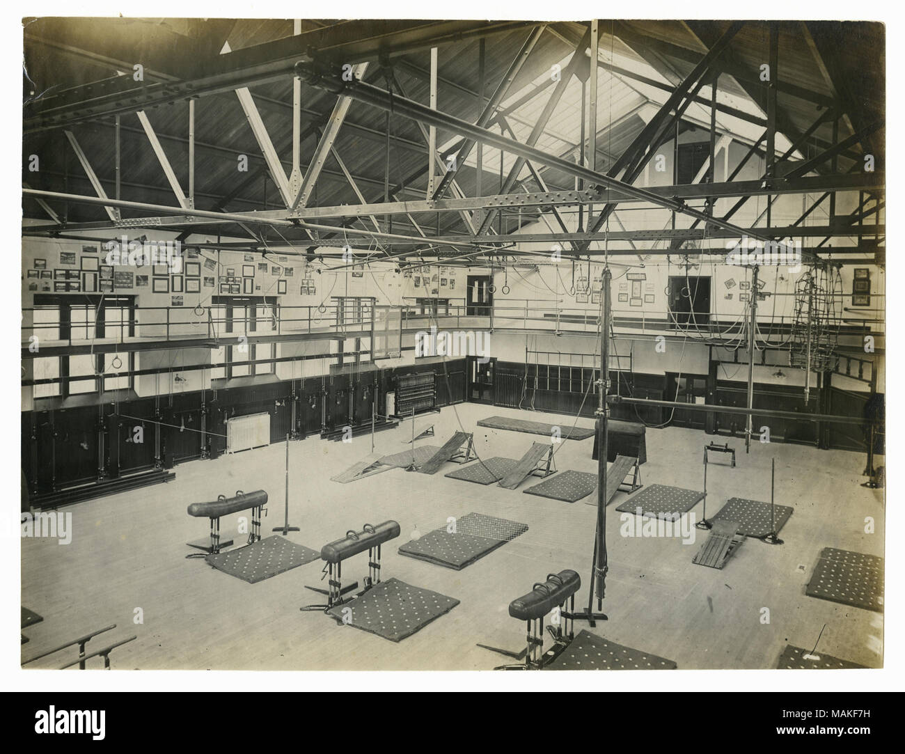 Horizontal photograph of a large interior space for a gym which includes a lot of gymnastic equipment such as pommel horses and rings as well as nets. There is a running rack around the top half of the gym. Title: 'A.G. Spalding and Bros. exhibit of a model gymnasium. Looking east from the running Track, showing a new model and latest improved horizontal bar.'  . 1904. Stock Photo