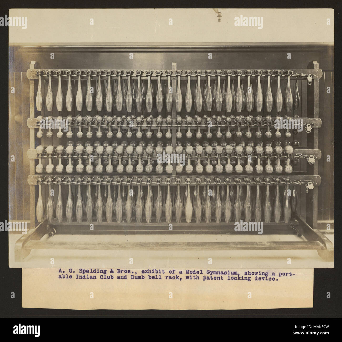 Horizontal photograph of a dumbbell rack with four rows of dumbbells in various sizes and two shapes. Title: 'A. G. Spalding and Bros., exhibit of a Model Gymnasium, showing a port-able Indian Club and Dumb bell rack, with patent locking device.'  . 1904. Stock Photo