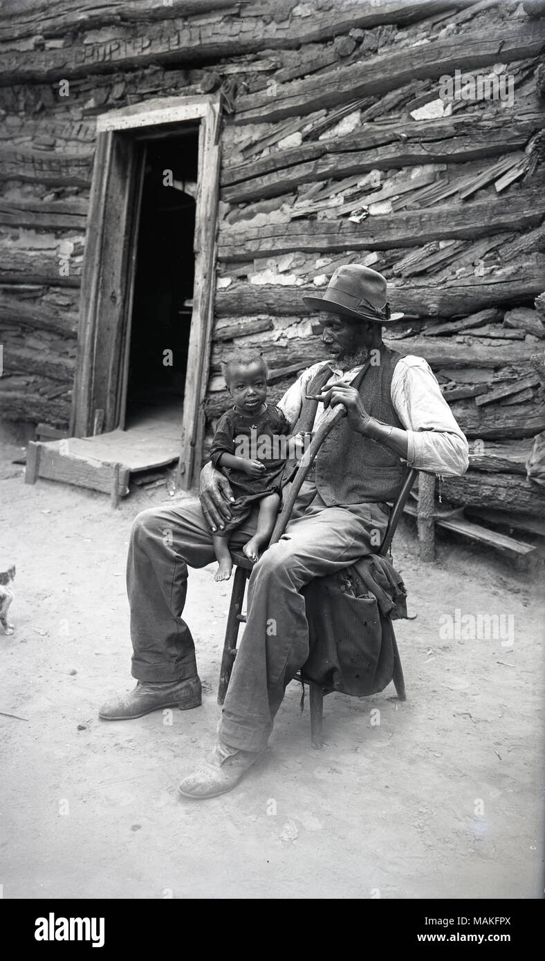 Vertical, black and white view of an elderly man, leaning on his cane, sitting in front of a log cabin with a small child on his lap. There is an open door way behind them. The child appears to have rickets or a leg deformity. Title: Robert Goodin, Vigus, Missouri, St. Louis County.  . circa 1910. Oscar Kuehn Stock Photo