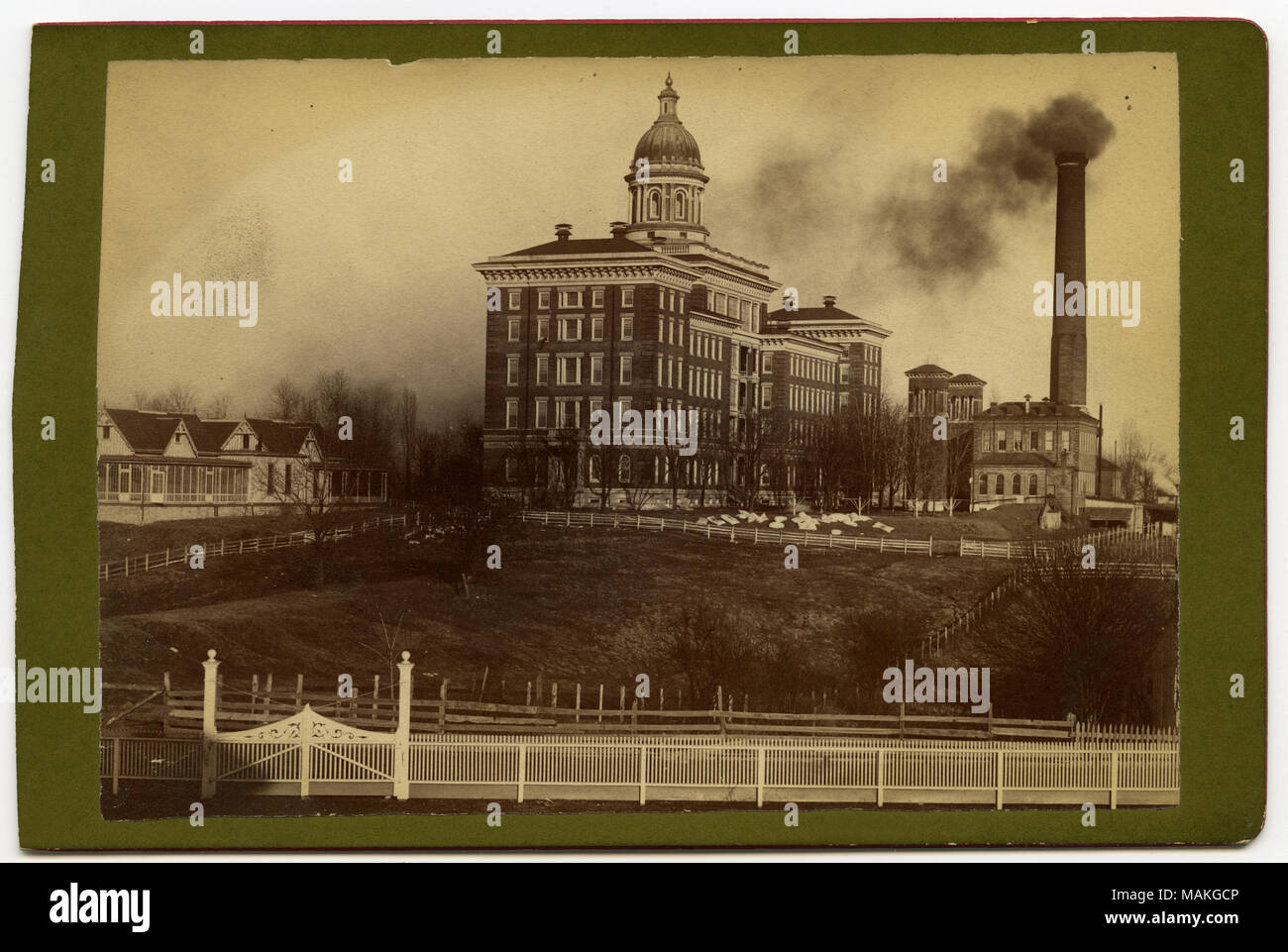 Horizontal, sepia photograph showing a view of a seven-storey brick complex with a small dome on top, a series of smaller outbuildings, and a large brick stack chimney with smoke at the far right. There are a series of fences around the grounds with a white one running along the front of the picture in the foreground. Handwritten note on the back reads: 'The Sanitarium formerly 'The Insane Asylum' 1890' Title: St. Louis Insane Asylum.  . 1890. Stock Photo