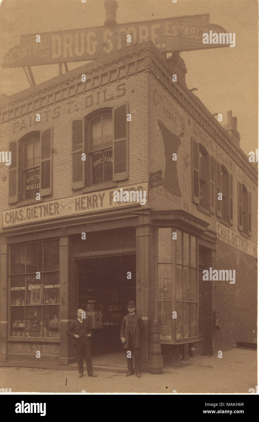 Vertical, sepia photograph of two men in suits standing in front of the open doorway of a drug store. Title: Charles Gietner Drugs, northwest corner of Seventh and Spruce Streets.  . circa 1870. Stock Photo