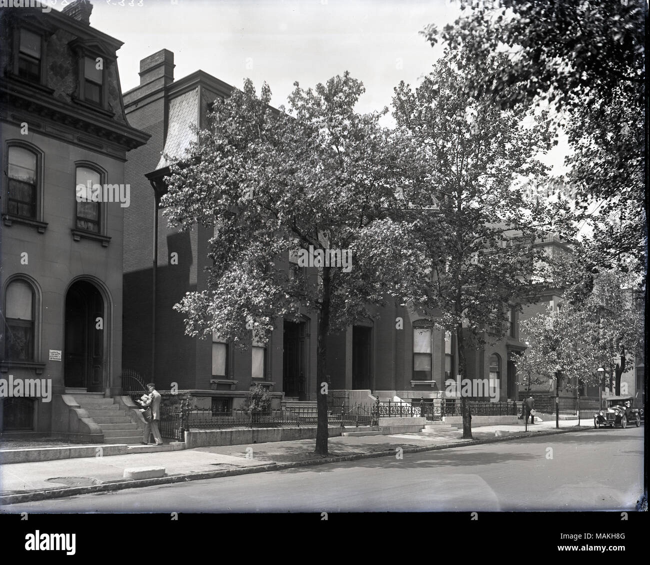 Horizontal, black and white photograph showing a view of three story houses along a tree-lined street. A mailman is delivering mail to the house on the left side of the print, and there is a car parked along the road on the far right side. Title: Street in Lafayette Square.  . 1908. Holt, Charles Clement, 1866-1925 Stock Photo