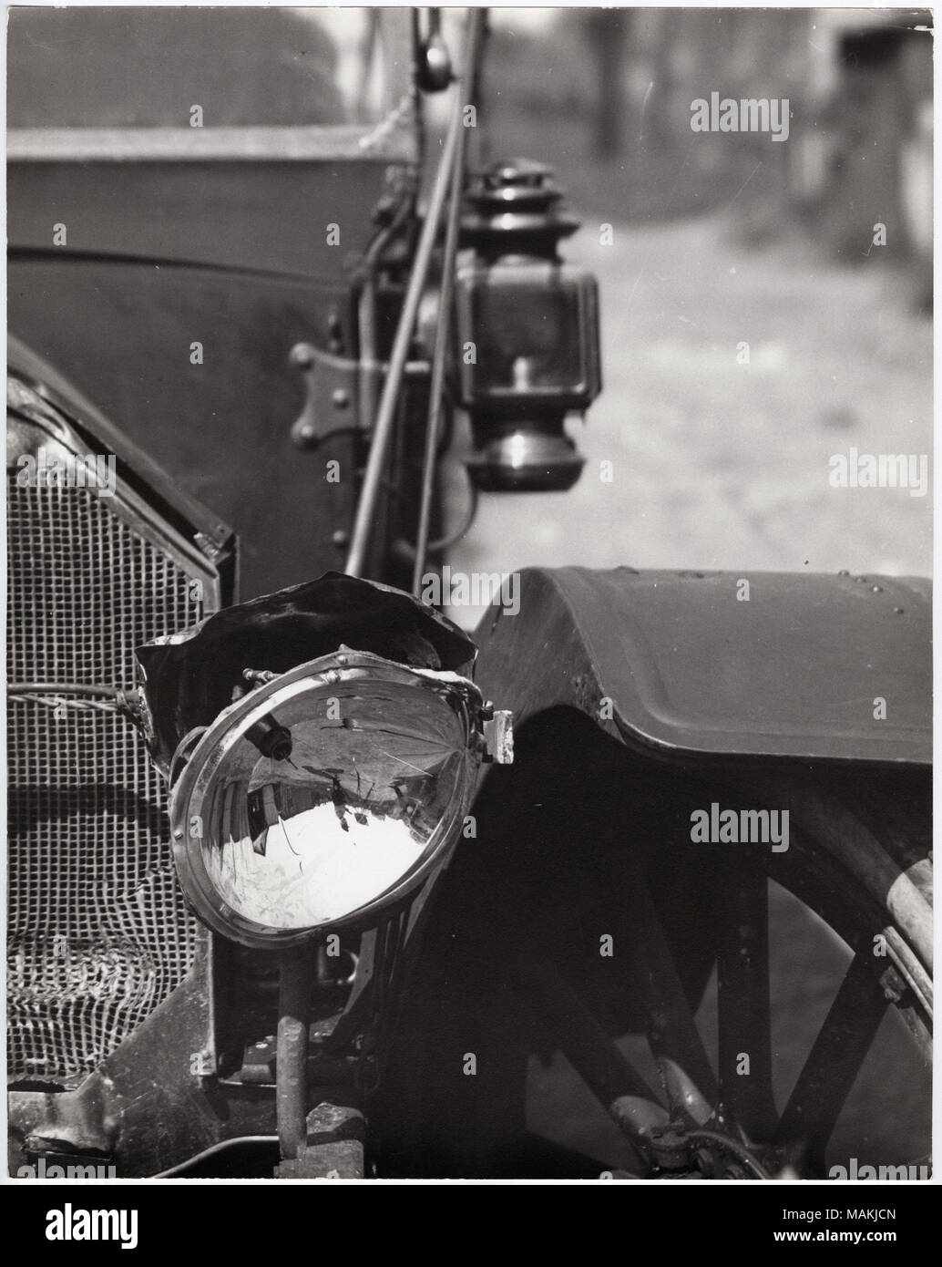 Vertical, black and white photograph showing a close-up view of a damaged automobile headlight and grille. The car's right headlight has been knocked out of place, and the grille has a dent in the lower right corner. The photographer with his camera and tripod can be seen reflected in the headlight. For the full image, see P0054-00025. Title: Close-up of a damaged automobile headlight.  . circa 1910. Holt, Charles Clement, 1866-1925 Stock Photo