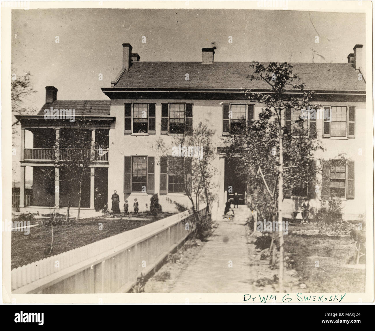 Horizontal, black and white photograph showing the Dr. John Gano Bryan residence at 1511 North Grand. The two-story house has an addition on the side, with a large balcony and porch area. Several adults and children are lined up in front of the house next to the new addition, while two children sit on the steps to the front door. These individuals are possibly members of the Huetteman and Uhlemeyer families, who may have owned or been living in different sections of the house at the same time. See the associated manuscript (ID# 6676) prepared by Dr. Swekosky for a detailed history of Bryan and Stock Photo