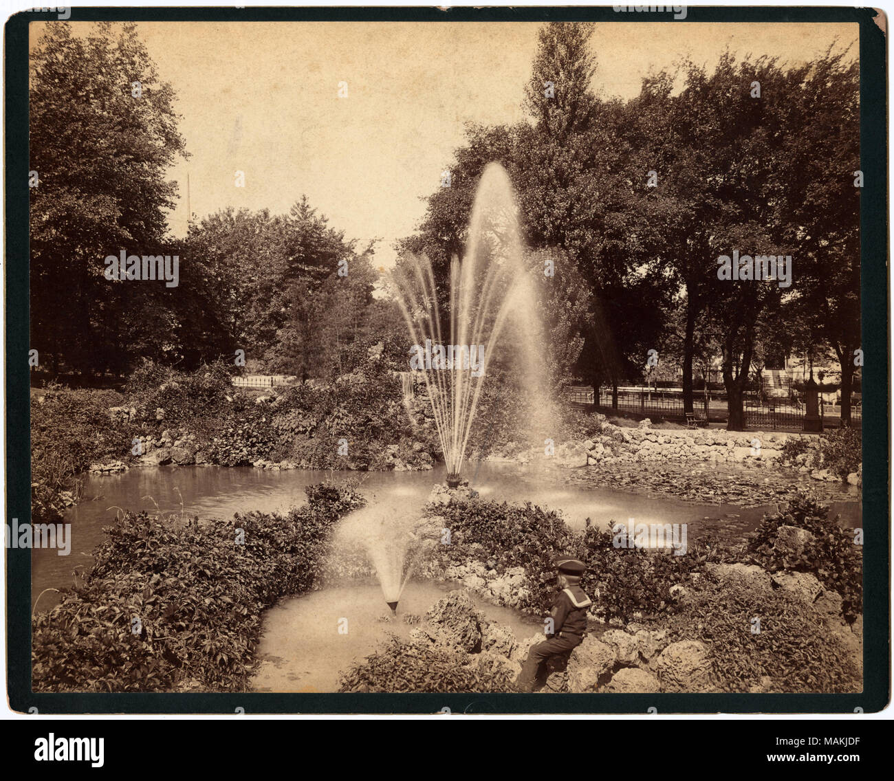 Horizontal, sepia photograph showing the rock garden at Lafayette Park. Rocks and foliage line the edges of a small pond with fountains in the center. A small boy in a sailor suit watches the fountains from a perch among the rocks. Large trees and a wrought-iron fence can be seen in the background. The back of the print has stamps from photographer Robert E. M. Bain and the St. Louis Globe Democrat. The Globe stamp indicates that the photograph appeared in the Gravure Section on December 4th, although there is no year given. A handwritten note, probably from Dr. William G. Swekosky, reads: 'Ra Stock Photo