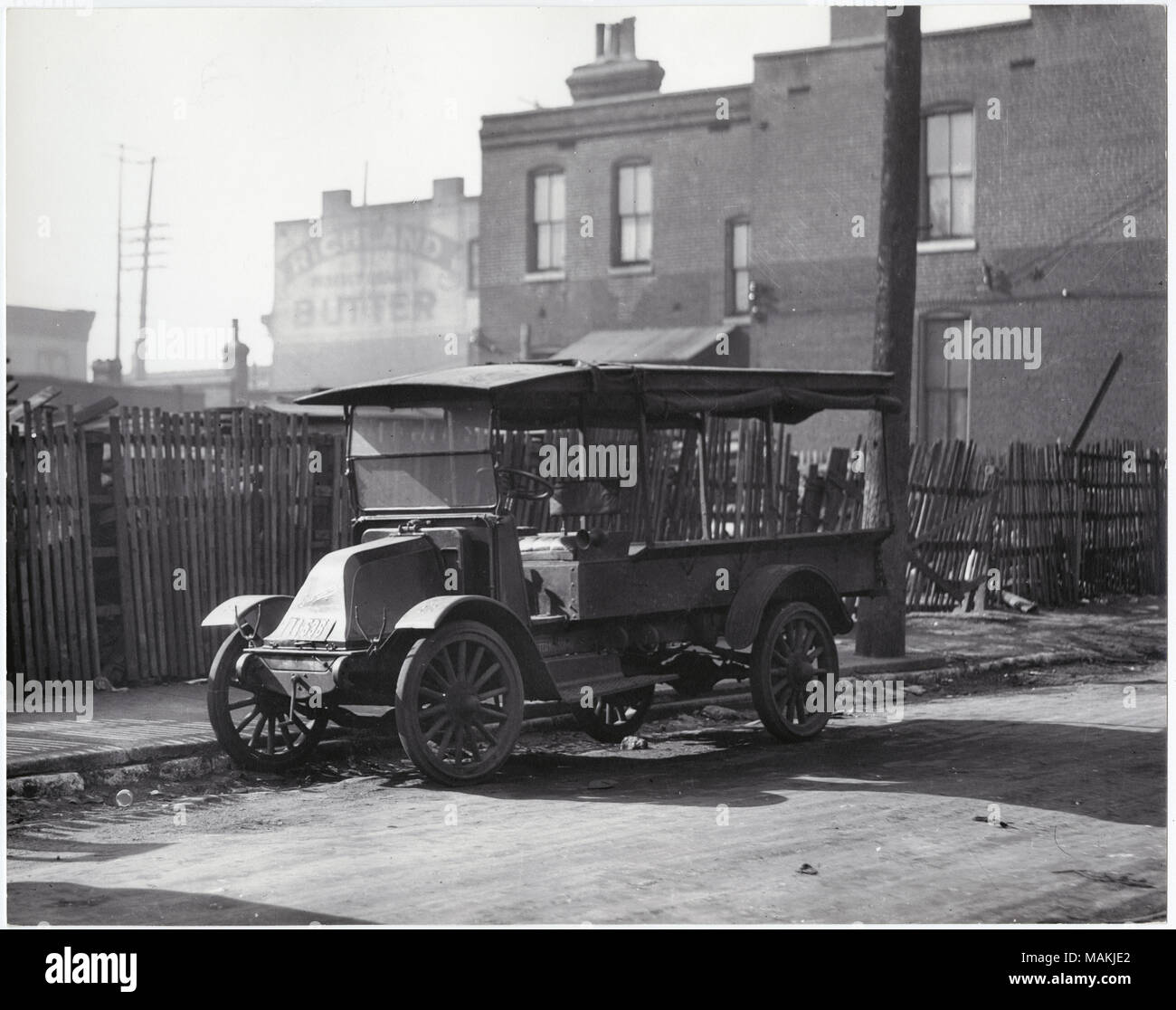 Horizontal, black and white photograph showing an open-air truck. The vehicle has an open cargo area behind the driver's seat. Although the body of the vehicle is open, a roof covers both the driver's seat and the cargo area. Rolled-up side curtains are visible along the roof line, and a hand-crank can be seen at the front of the vehicle. A wooden fence and several unidentified buildings, one with a large advertisement for Richland butter, are in the background. Title: Open-air truck, unidentified location.  . circa 1915. Holt, Charles Clement, 1866-1925 Stock Photo