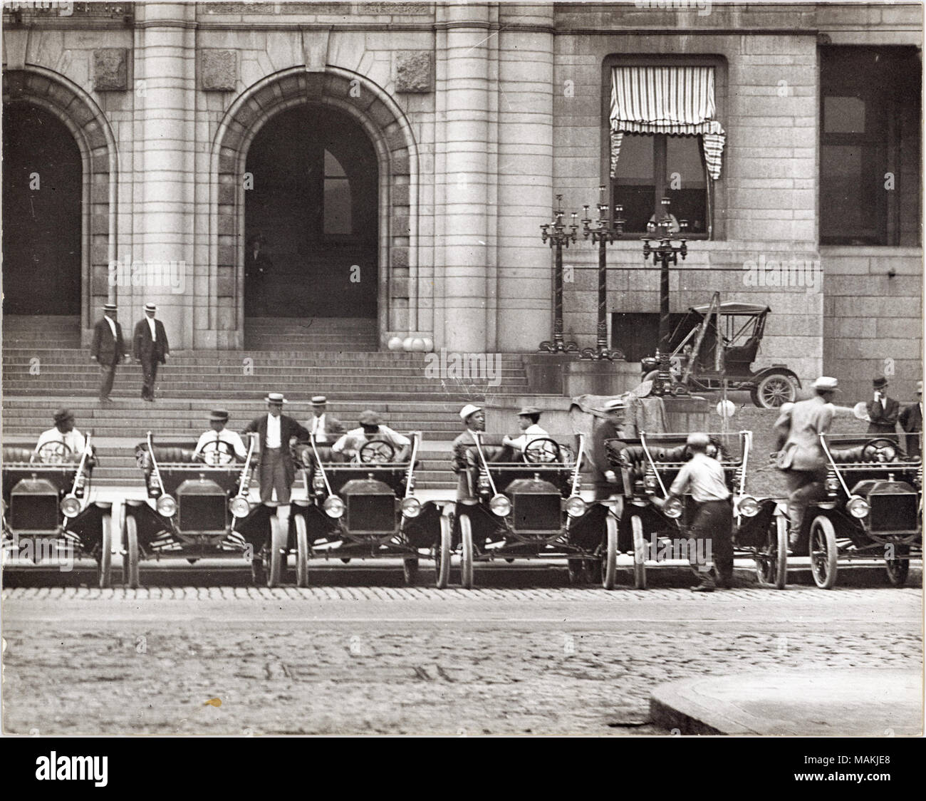 Horizontal, black and white photograph showing a row of six Model T Fords parked in front of St. Louis City Hall, probably near the intersection of 12th Street, now Tucker Boulevard, and Walnut Street . Men are seated in the driver's seat of four of the cars, and one man climbs in or out of a fifth car. Other men can be seen walking on the sidewalk in the background. A typed note on the back of the print, possibly an exhibit caption, reads: 'Model T Fords, ca. 1909. Fourteen of these automobiles were bought by City Hall. The cars had a 4 cylinder engine and 2 speed transmission, features which Stock Photo
