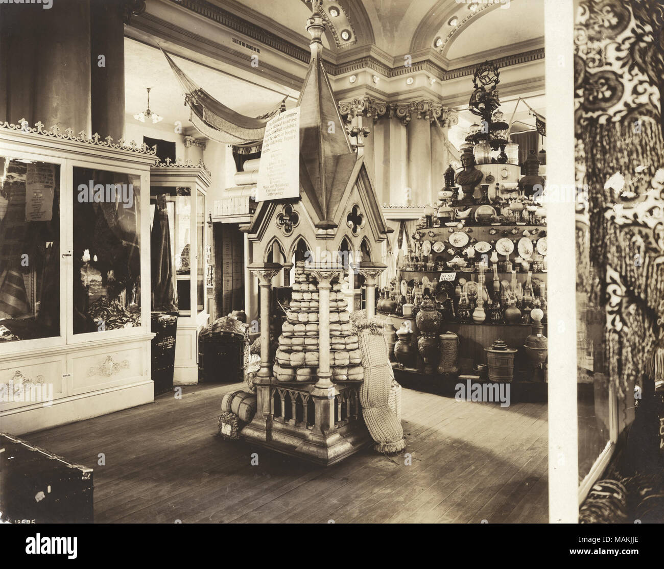Horizontal, sepia photograph showing several product displays. In the center of the photograph is a display built to look like a small building with a steeple-like roof. To the right of that display is a tiered circular case featuring sculpture, plates and pottery. Title: Interior of Brazil's exhibit in the Palace of Manufactures at the 1904 World's Fair.  . 1904. Official Photographic Company Stock Photo