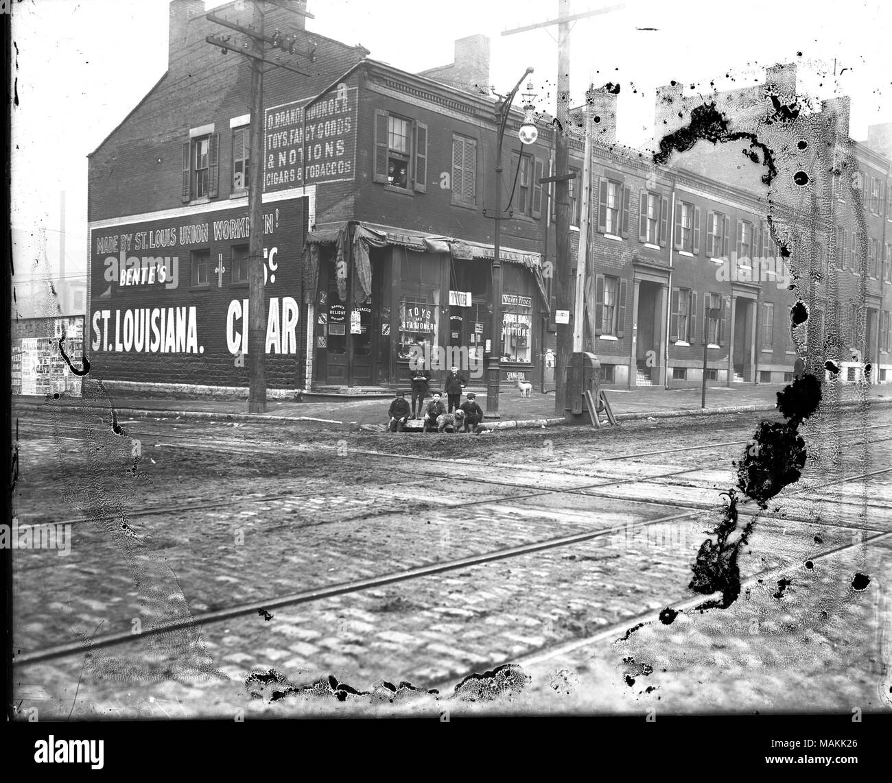 Horizontal, black and white photograph showing a view of two story row buildings from the opposite corner of an intersection. The building on the corner is a toy and notions store, and a small group of boys are on the sidewalk at the corner. Signboards painted on the side of the building read: 'O. Brandenburg. Toys, Fancy Goods & Notions. Cigars & Tobaccos' and 'Made by St. Louis Union Workmen! Bente's St. Louisiana. Cigar.' Title: Children in front of O. Brandenburger Toy, Fancy Goods, and Notions store.  . between 1901 and 1909. Holt, Charles Clement, 1866-1925 Stock Photo