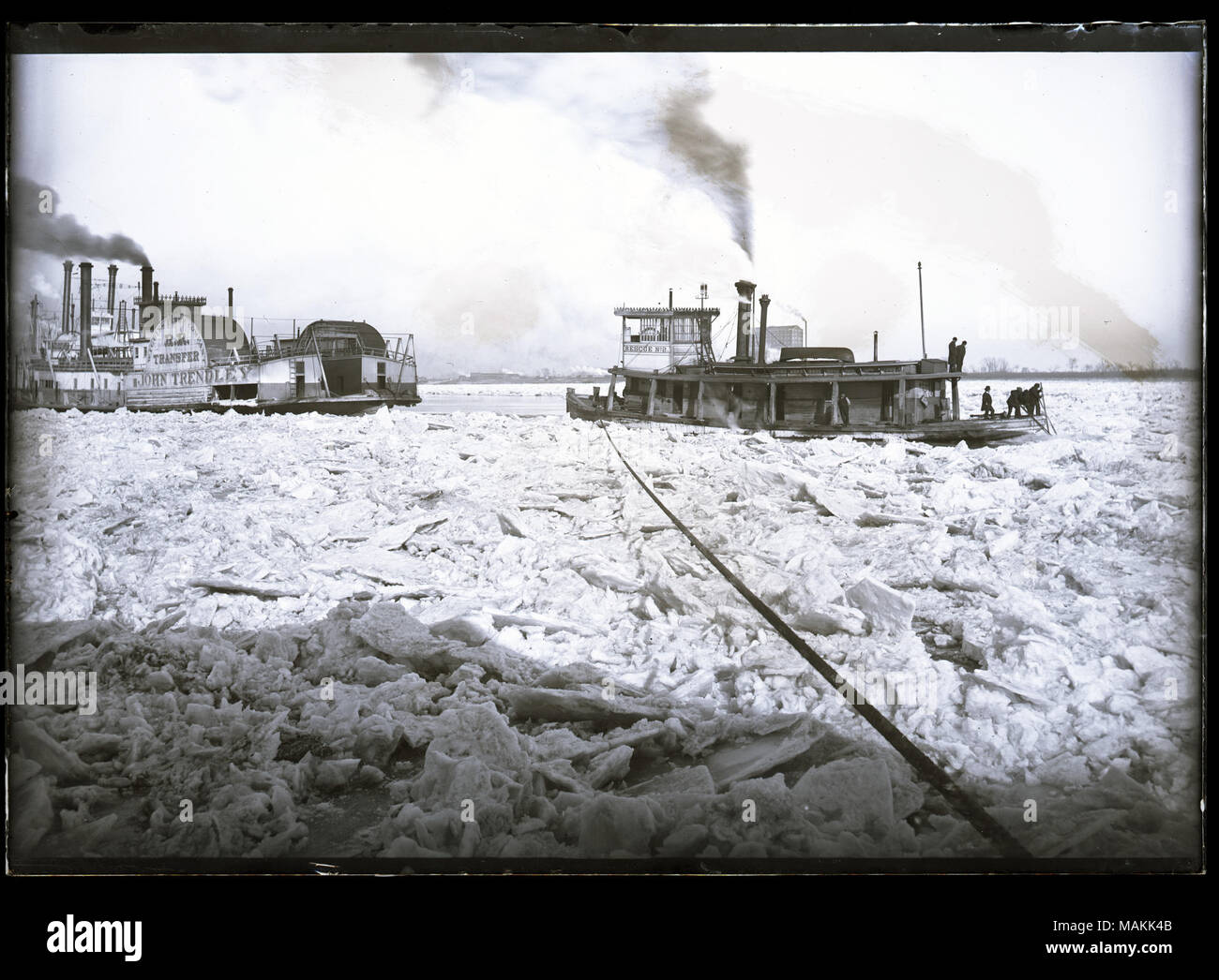 Horizontal, black and white photograph showing steamboats caught on an icy river, possibly during the ice gorge of 1887. The steamboat John Trendley, owned by the Wiggins Ferry Company, is on the left, with an unidentified steamboat behind it. These steamboats appear to be trapped in the ice. On the right, there is a boat named Rescue No. 2, with a rope running from the boat's deck into the foreground. Several men are standing on the boat's deck, using poles or other long tools. The path in front of Rescue No. 2 appears blocked by ice, and there are ice chunks throughout the foreground. Title: Stock Photo