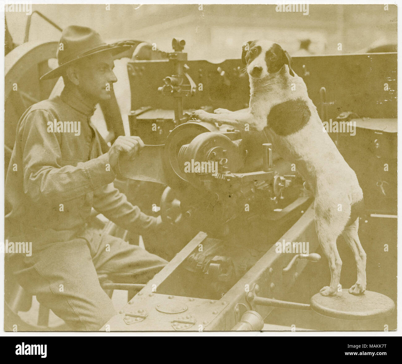 Horizontal, sepia photograph showing a uniformed soldier working on a large piece of machinery. A dog is bracing its front paws on part of the machinery while looking toward the photographer. Title: Soldier Working on Large Machinery With a Dog Standing Nearby.  . between circa 1914 and circa 1918. Michel, Carl Stock Photo