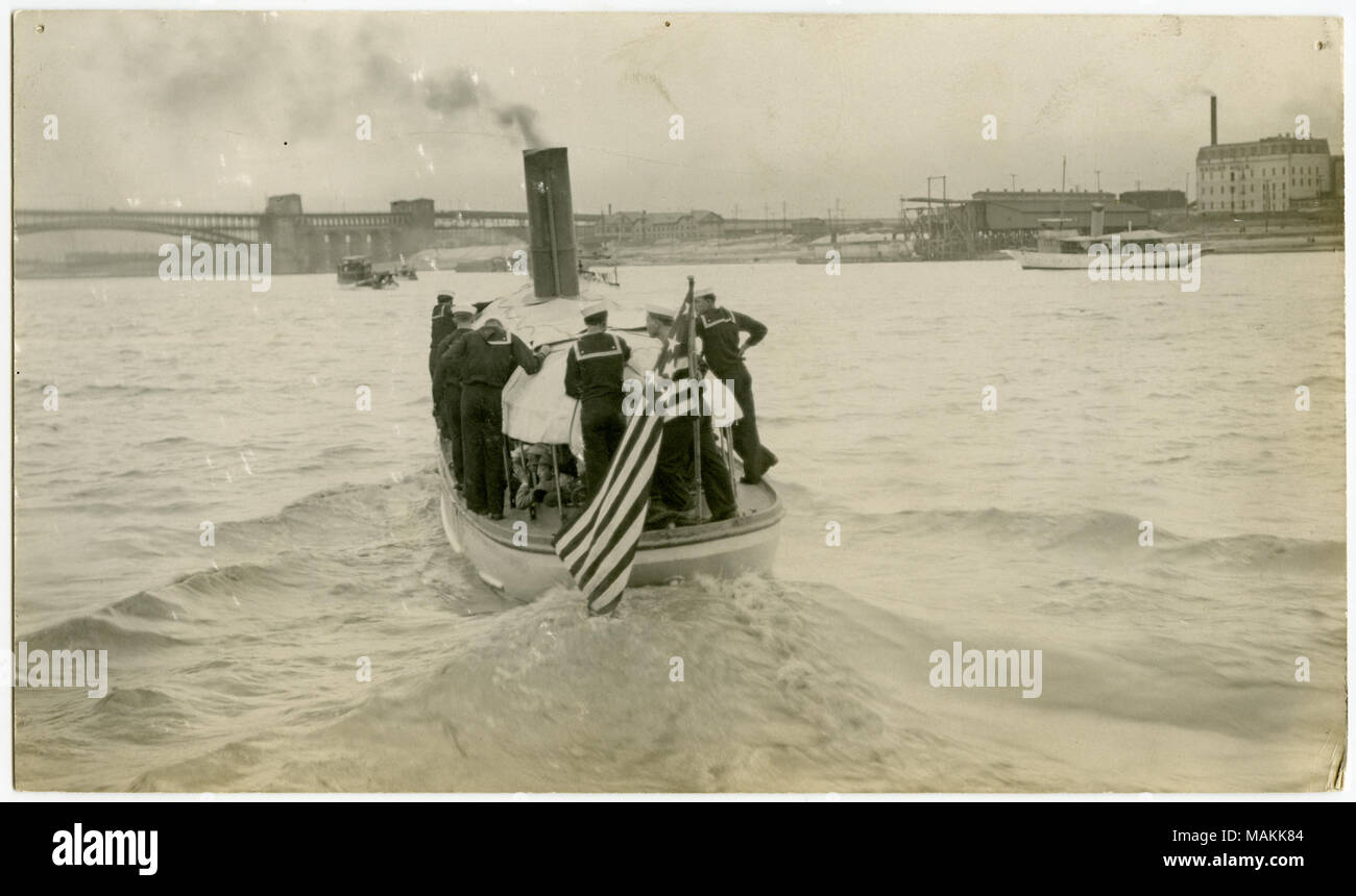 Horizontal, sepia photograph showing a steamboat with an American flag flying from the back of the boat moving along the Mississippi River. Some of the pasengers inside the boat are wearing civilian clothes while others are in Navy uniforms. Sailors in Navy uniforms are standing along the edges of the boat. The Eads Bridge is visible in the distance. Title: Sailors and Civilians Riding in a Steamboat Down the Mississippi River.  . between circa 1914 and circa 1918. Michel, Carl Stock Photo
