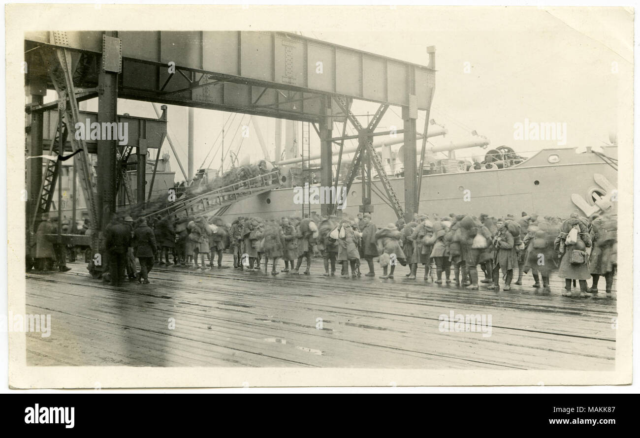 Horizontal, black and white photograph of a line of men in military uniforms, holding bags on a dock, and preparing to board a ship on April 14, 1919. The photograph is located in the St. Louis 12th Engineer's Regiment album, on pg. 39. Title: 12th Engineer Regiment, Embarking on the S.S. Cape May, Bassens, France.  . April 1919. Stock Photo