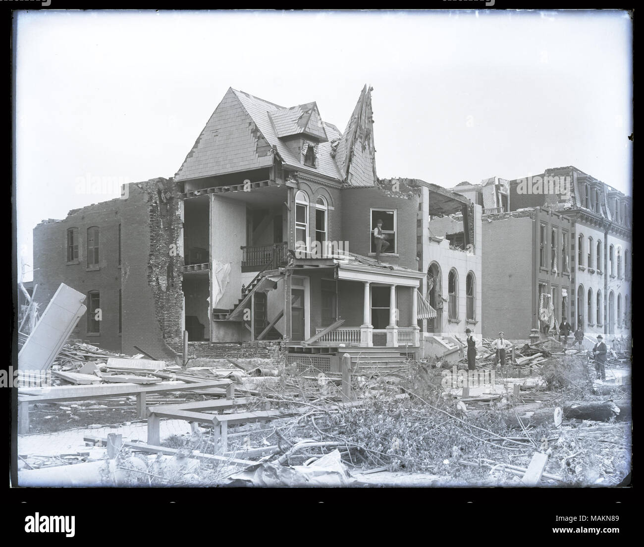 Horizontal, black and white photograph showing destruction in the Lafayette Park neighborhood after a tornado, probably the 1896 tornado. Photograph was probably taken at the corner of Park Avenue and Mississippi Avenue. Five houses have had parts of their roofs, walls, and windows destroyed. An interior staircase of one home can be seen. Piles of rubble and fallen trees can be seen throughout the photograph. Several men are standing amid rubble on the right. Title: Destruction in the Lafayette Park neighborhood after tornado, probably the 1896 tornado.  . 1896. Stock Photo
