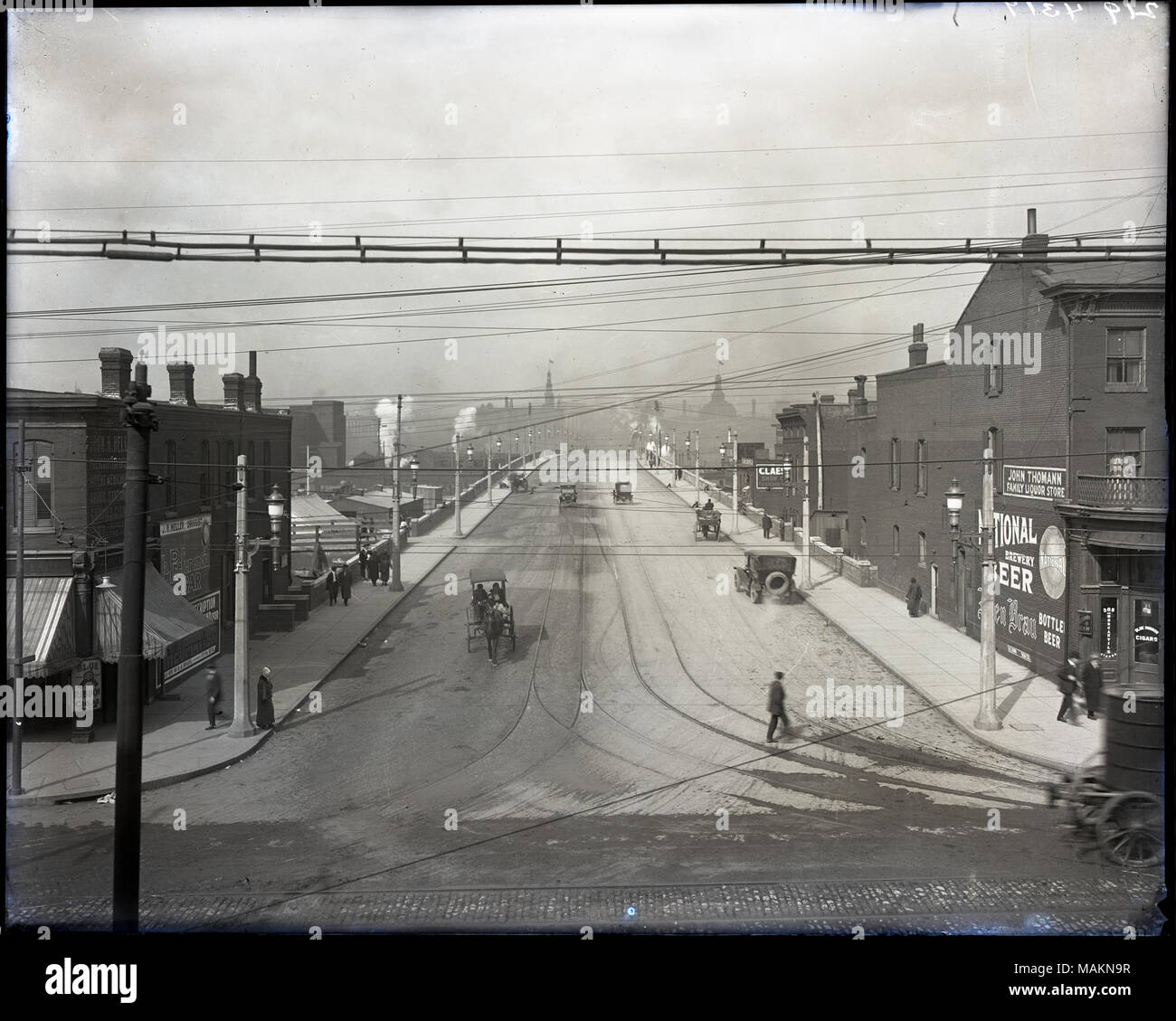 Horizontal, black and white photograph showing a bird's eye view of the intersection of 12th Street and Chouteau. The view looks down 12th Street, later renamed Tucker Boulevard. Several horse drawn wagons and buggies and several cars are driving on 12th Street, which is lightly covered with snow. Pedestrians are walking down the sidewalk and crossing the street. J. H. Heller, druggist, is on the left, probably at 1201 Chouteau. John Thomann, family liquor store, is on the right, probably at 1113 or 1114 Chouteau. Title: Bird's eye view of the intersection of 12th Street and Chouteau.  . betwe Stock Photo