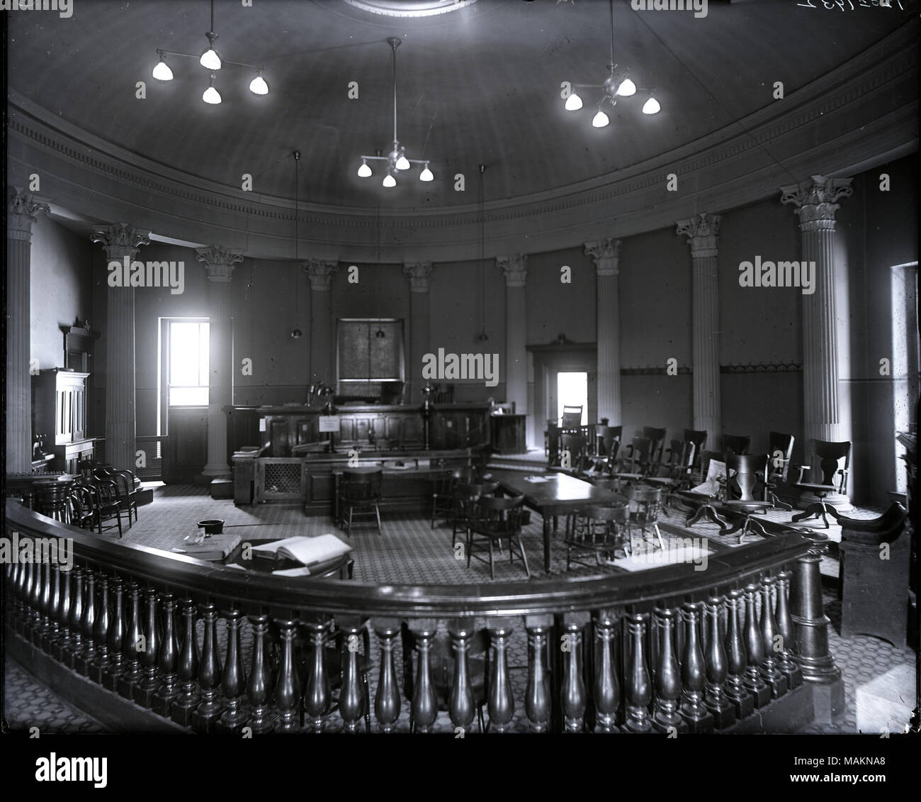 Horizontal, black and white photograph showing an interior view of the court room at the Old Courthouse. A wooden rail curves around the room in the foreground, and the judge's bench can be seen in the background. Columns line the walls of the room, which also has a domed ceiling. Title: Interior view of the court room at the Old Courthouse.  . between 1900 and 1920. Stock Photo