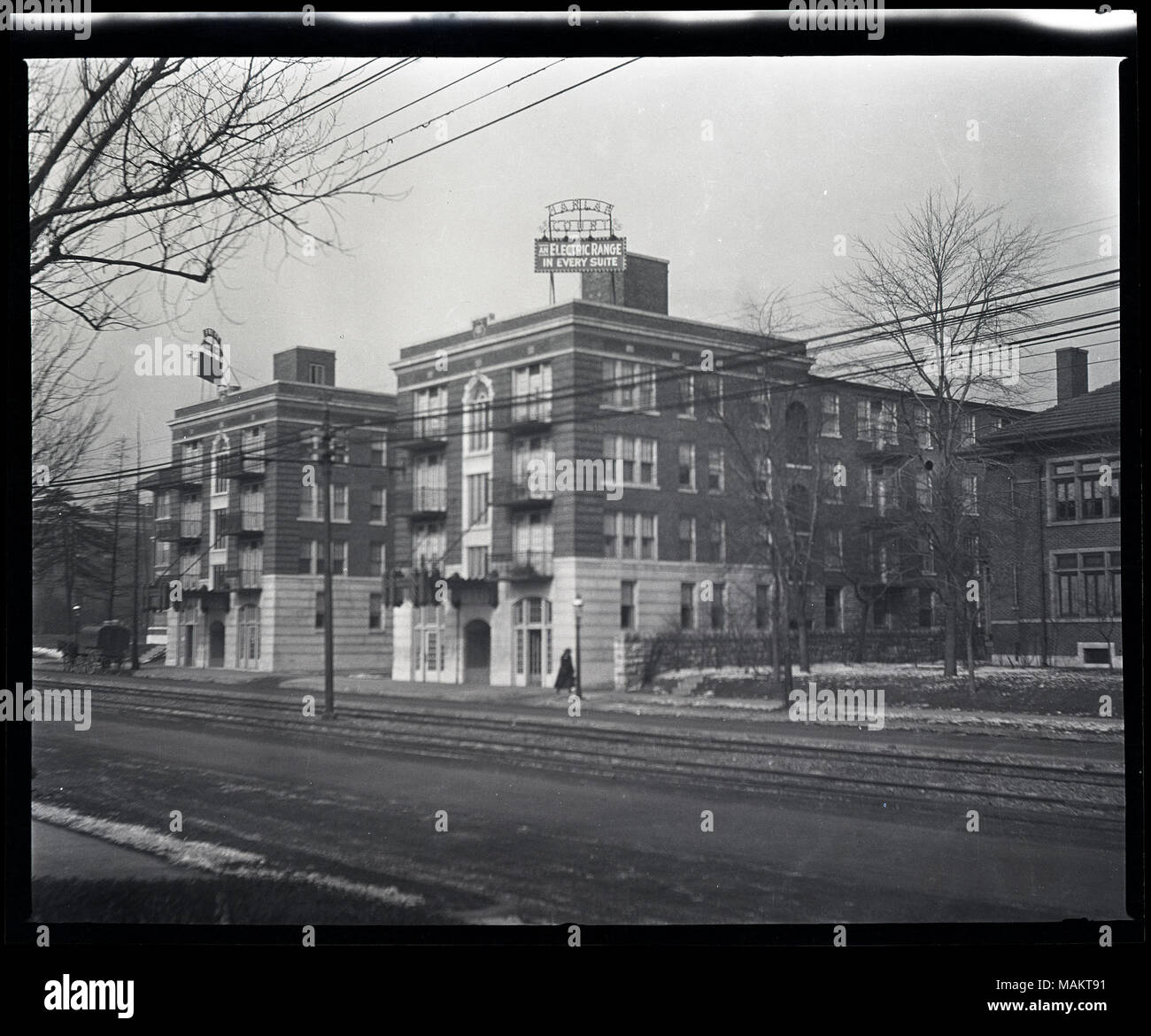Horizontal, black and white photograph showing the two wings of the Harlan Court apartment building located at 5457 Delmar Boulevard. The apartment building has two four-story wings built of brick and stone. A sign that reads 'Harlan Court/An Electric Range in Every Suite' is on the roof of each wing. Metal balconies and a metal canopy on the front facade of the building can also be seen. The view was taken at an angle from across the street and shows the streetcar rails in the middle of the road. A horse pulling a covered cart can be seen in the distance on the left. Title: Harlan Court Apart Stock Photo