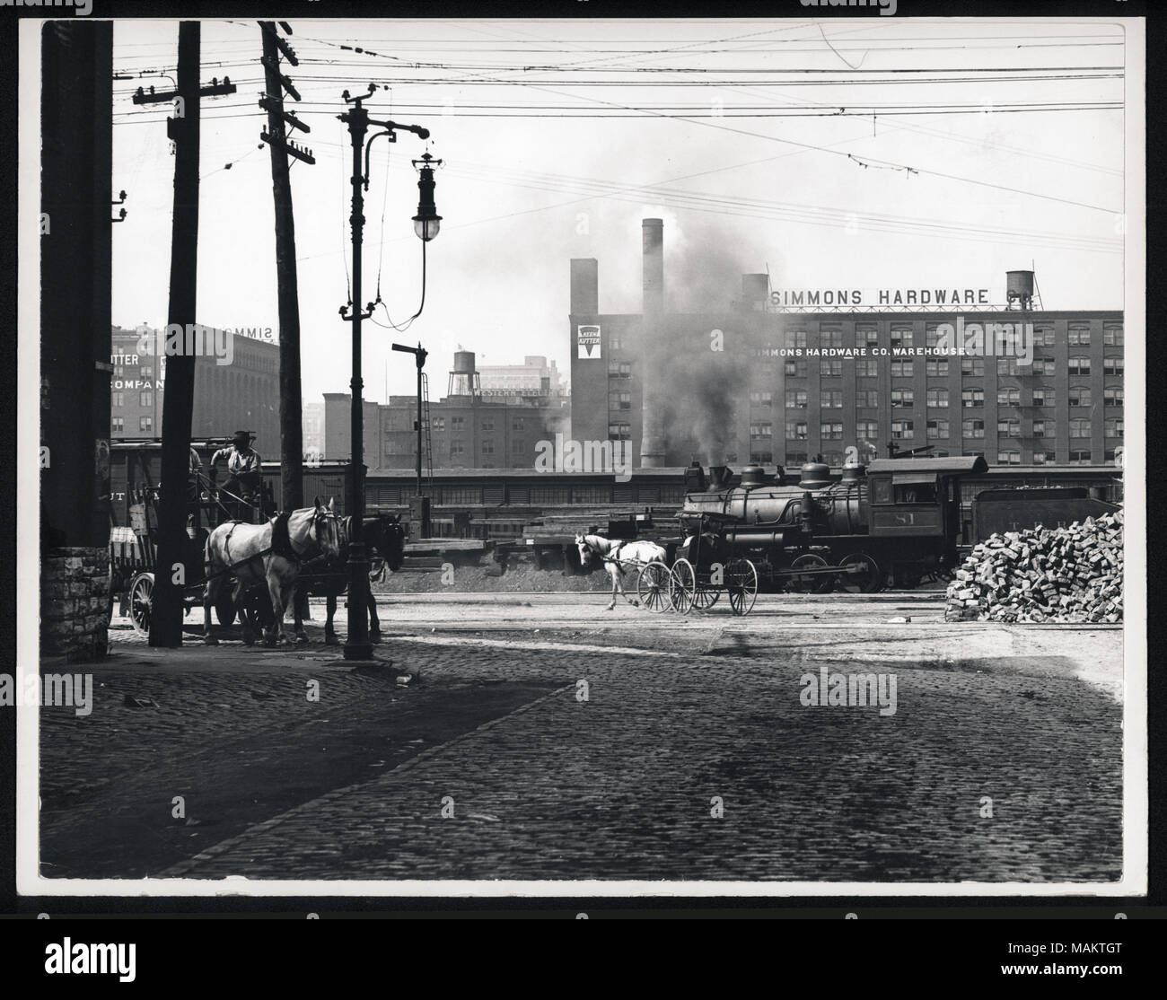 Horizontal, black and white photograph of a brick factory building for Simmons Hardware Co. Horse-drawn carriages and a train are at the front of the photograph. Title: Simmons Hardware seen from Eighth and Spruce.  . between circa 1800 and circa 1900. Swekosky, William G., 1895-1964 Stock Photo