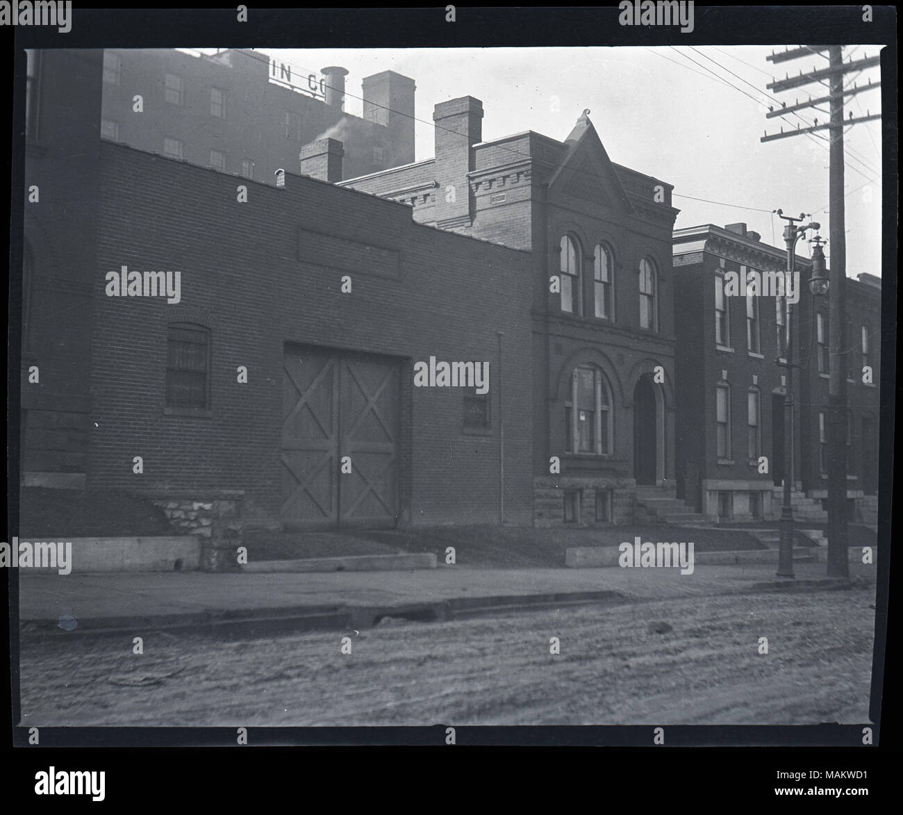 Horizontal, black and white photograph showing a row of two-story brick residential buildings next door to an unidentified industrial or commercial building. The residential buildings have decorative brickwork along the roofline and around the windows. It is unclear if the buildings are single family homes or multi-family apartment buildings. A small poster with a light background and a cross in the center is displayed in one of the windows, possibly a Red Cross Service Flag given out during World War I. The industrial or commercial building is also brick. There is a set of large double doors  Stock Photo