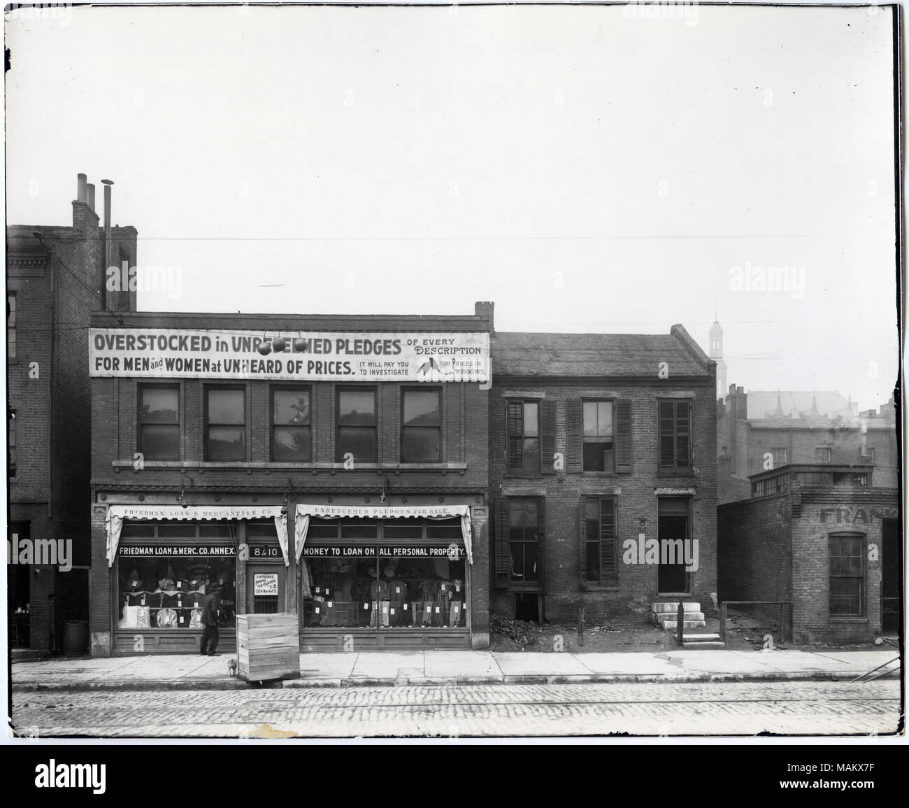 Horizontal, Black and white photograph of a two story brick commercial and residential building. Advertisements for Friedman Loan and Mercantile Company are placed at the top of the building and above the display windows. A decorative molding is above the display windows which showcase multiple items of clothing. A wooden crate is out front on the sidewalk with a dog standing next to it and a man is on the sidewalk. The second building is a two story brick building with shutters around the windows. The road is paved of brick. Title: Fourteenth (8-10). Friedman Loan and Mercantile Company Annex Stock Photo