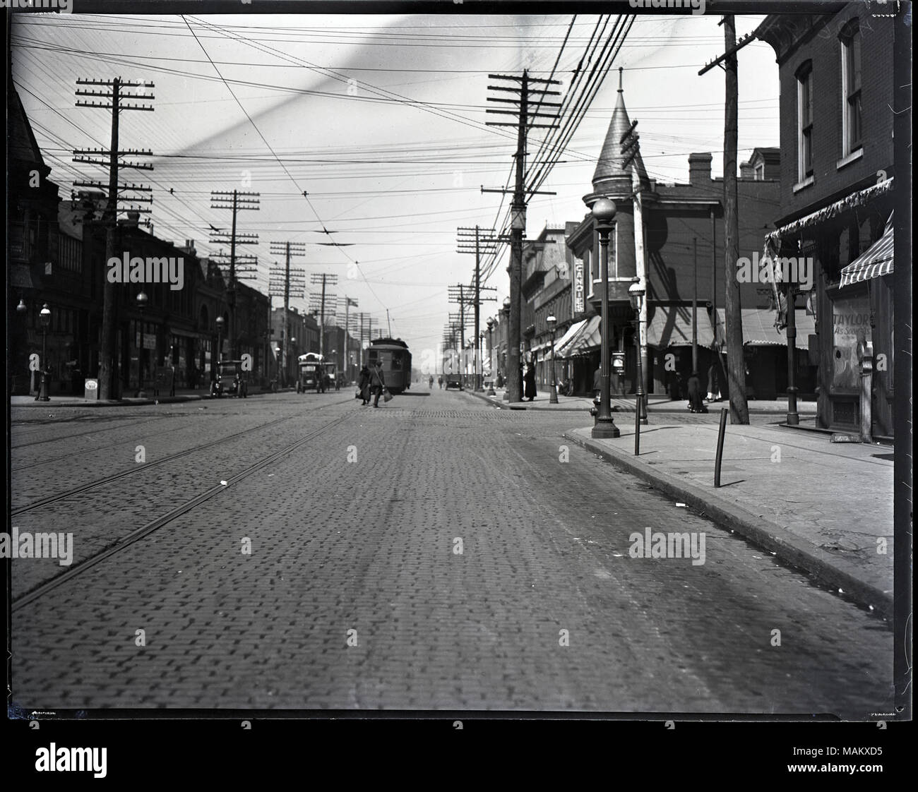 Horizontal, black and white photograph showing the view looking down the street at the intersection of Taylor and Easton. Easton was later renamed Dr. Martin Luther King Drive. A variety of stores and businesses are housed in the two-story brick buildings that line both sides of the cobblestone street. Streetcar tracks run down the center of the street, and a streetcar can be seen in the background. There are also several cars and pedestrians. A note along the upper edge on the back of the negative reads: '406-9702 / Taylor and Easton.' Title: View looking down the street at the intersection o Stock Photo