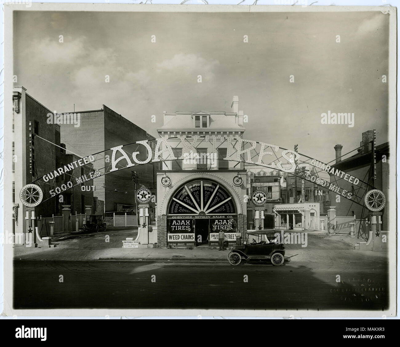 Horizontal, black and white photograph showing advertising for Ajax Tires at a Texaco gas station. Driveways wrap around both sides of a two-story building. The building has large advertisements for Ajax tires in the windows, and a sign for Guenther-Methudy Sales Co. over the door. A sign stretching across the driveways in front of the building has letters outlined in light bulbs and reads: 'Ajax Tires / Guaranteed 5000 miles.' Signs for Texaco Gasoline can be seen on either side of the main building, and on a small building in the background. Several cars are parked around the building. Title Stock Photo