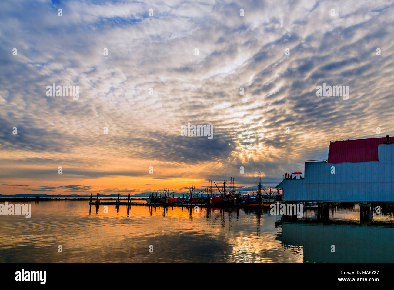 Sunset on the river near the pier of fishing ships with reflection in the water, huge cloudy sky and silhouettes of people under a canopy in a cafe Stock Photo