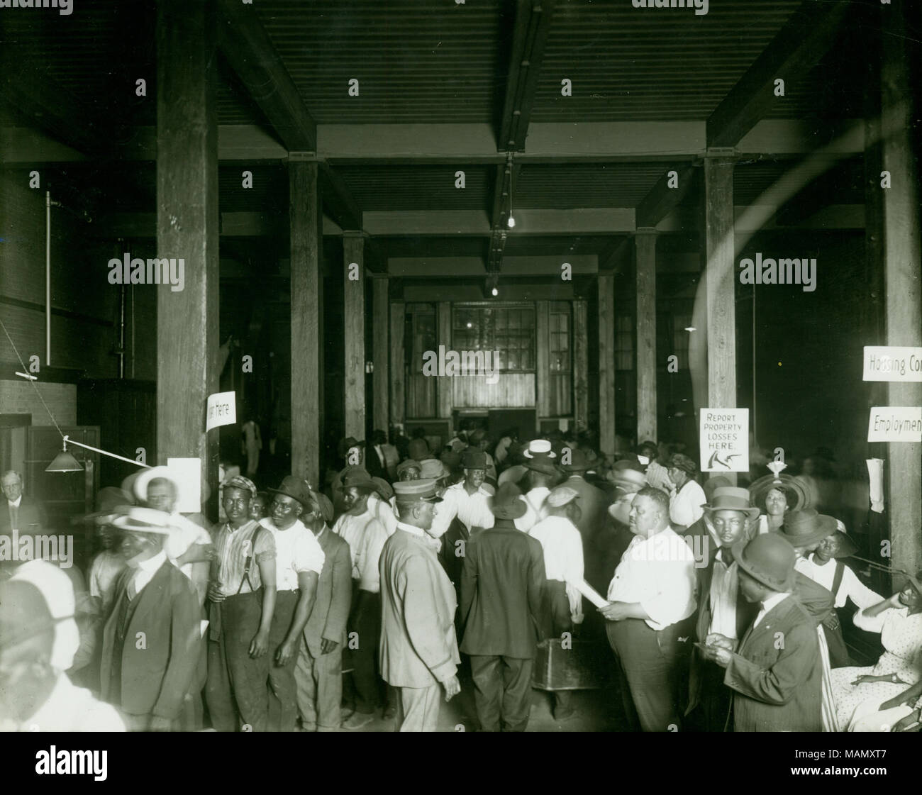 Horizontal, black and white photograph showing a large open room where a group of African Americans, mostly men, There is a large central aisle in the room. Along the left side men are standing in a line, in the center people are exiting the room, and along the right side people are seated or waiting. There are some signs posted on poles in the room reading 'Report Property Losses Here', 'Housing Con[ditions]', and 'Employment.' The photograph was possibly taken at the Municipal Lodging House (110 South 12th Street). Title: Group of African American men reporting property losses around time of Stock Photo