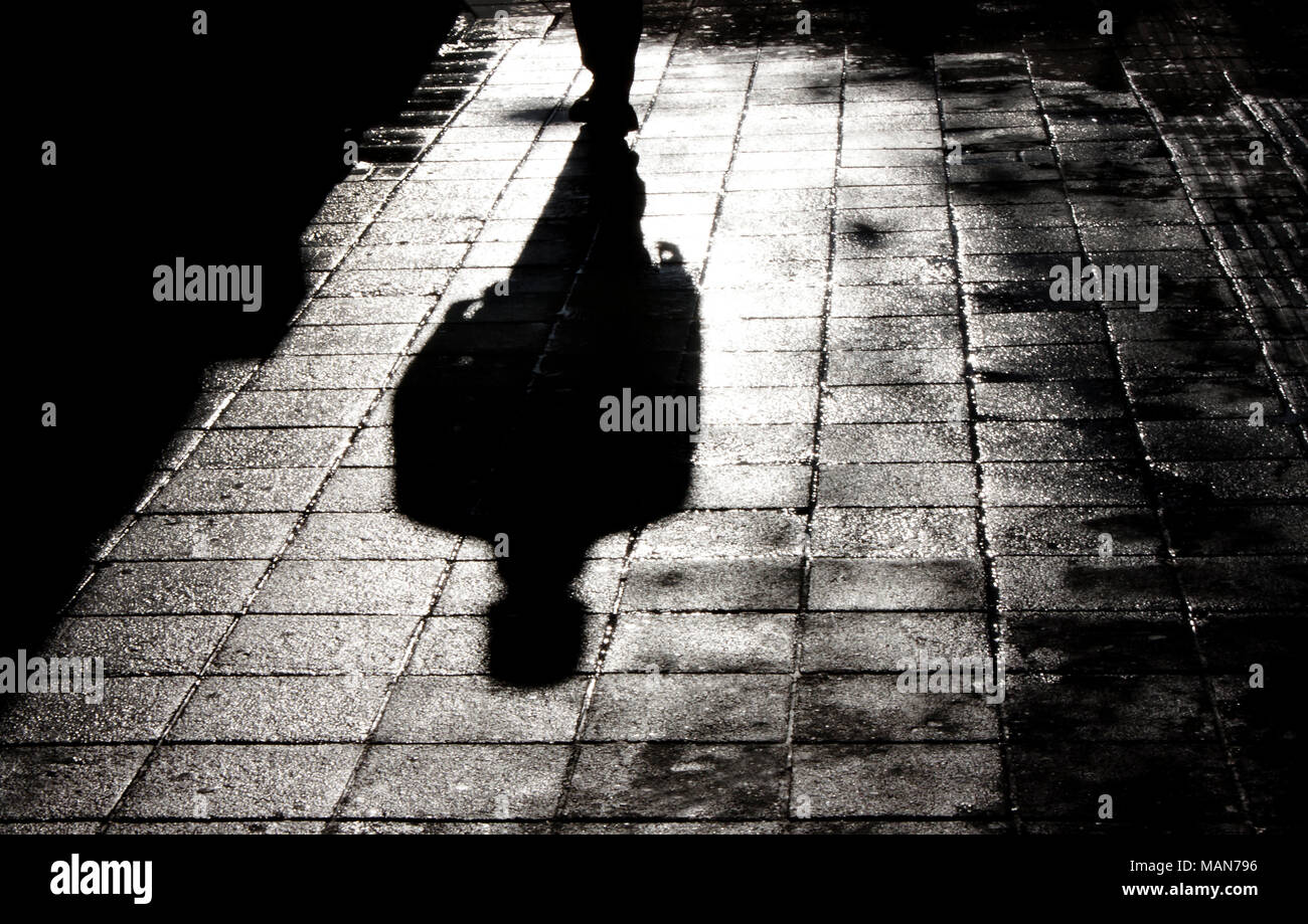 Blurry shadow and silhouette of a man standing in the night on wet city street sidewalk with water reflection in black and white Stock Photo