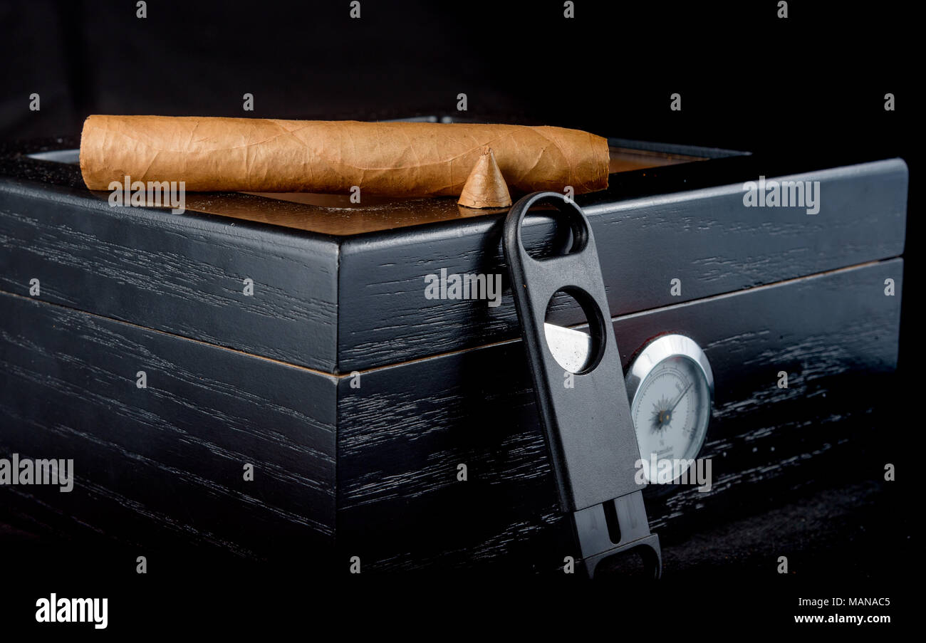 A rich smelling cigar that has been straight cut with smoke rising. Resting on a black humidor next to a straight cigar cutter. Black background Stock Photo