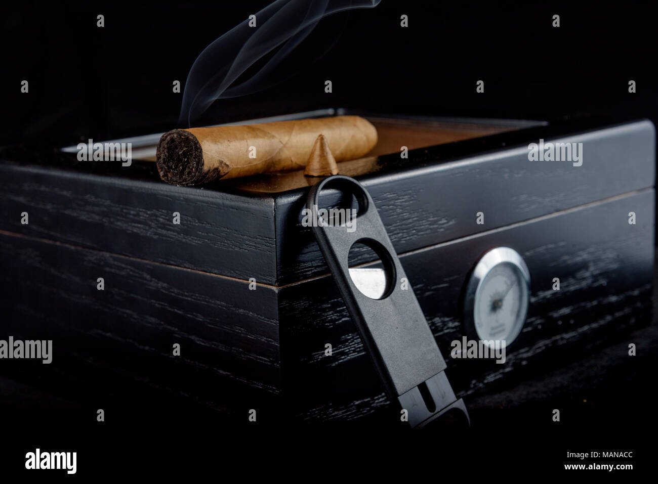 A rich smelling cigar that has been straight cut with smoke rising. Resting on a black humidor next to a straight cigar cutter. Black background Stock Photo