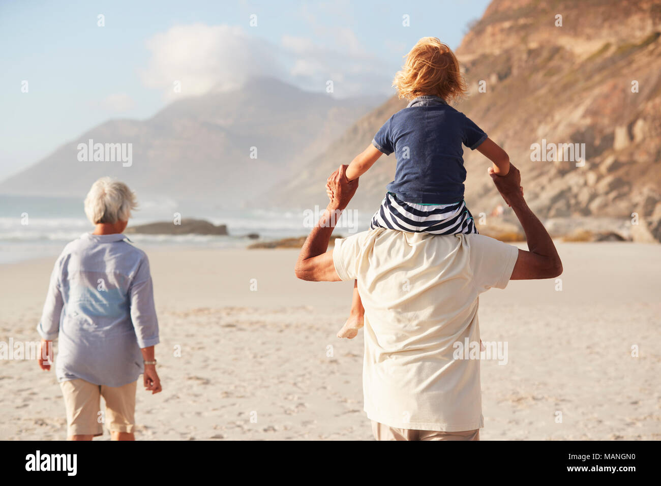 Grandparents Carrying Grandson On Shoulders On Walk Along Beach Stock Photo