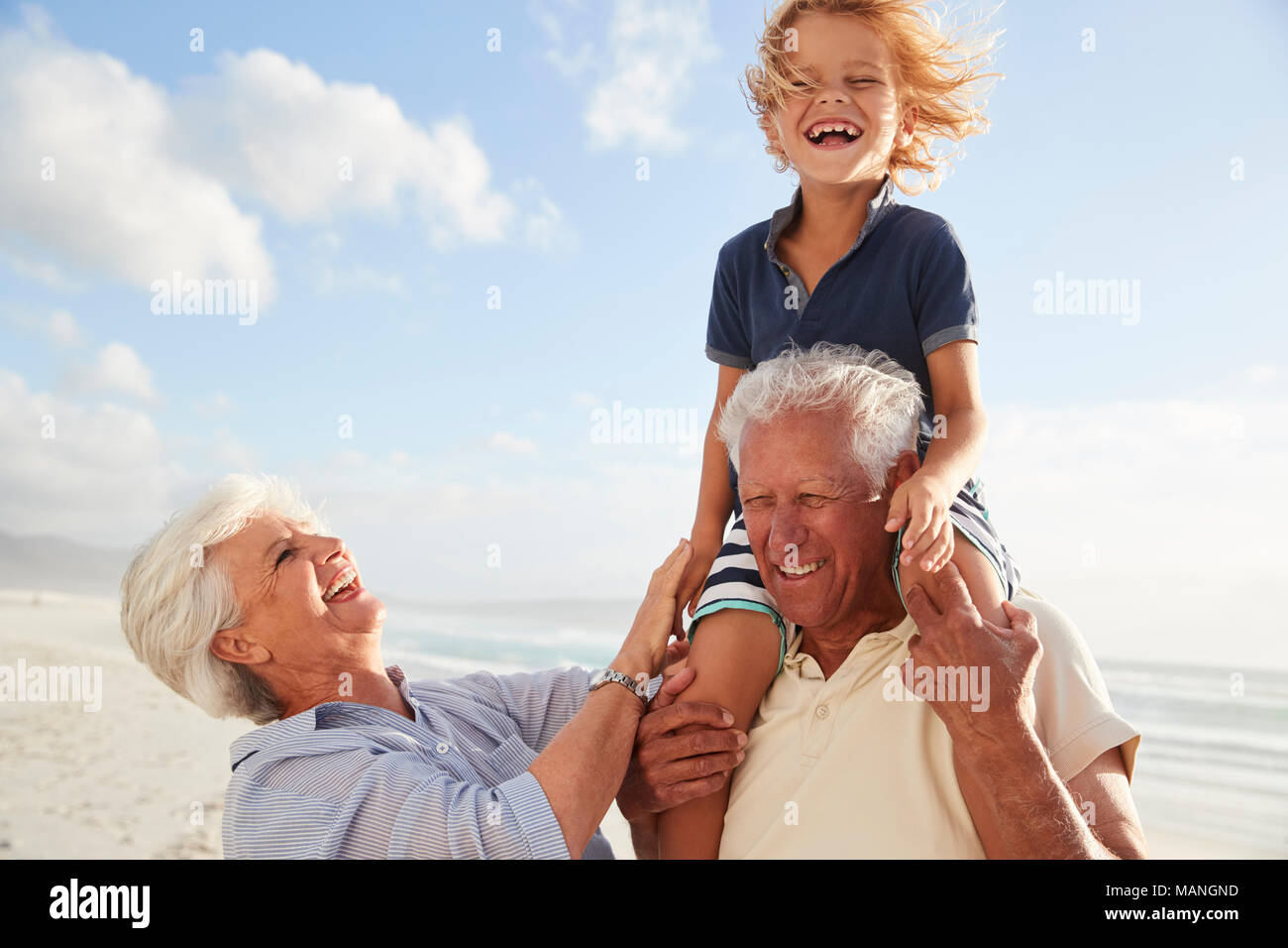 Grandparents Carrying Grandson On Shoulders On Walk Along Beach Stock Photo