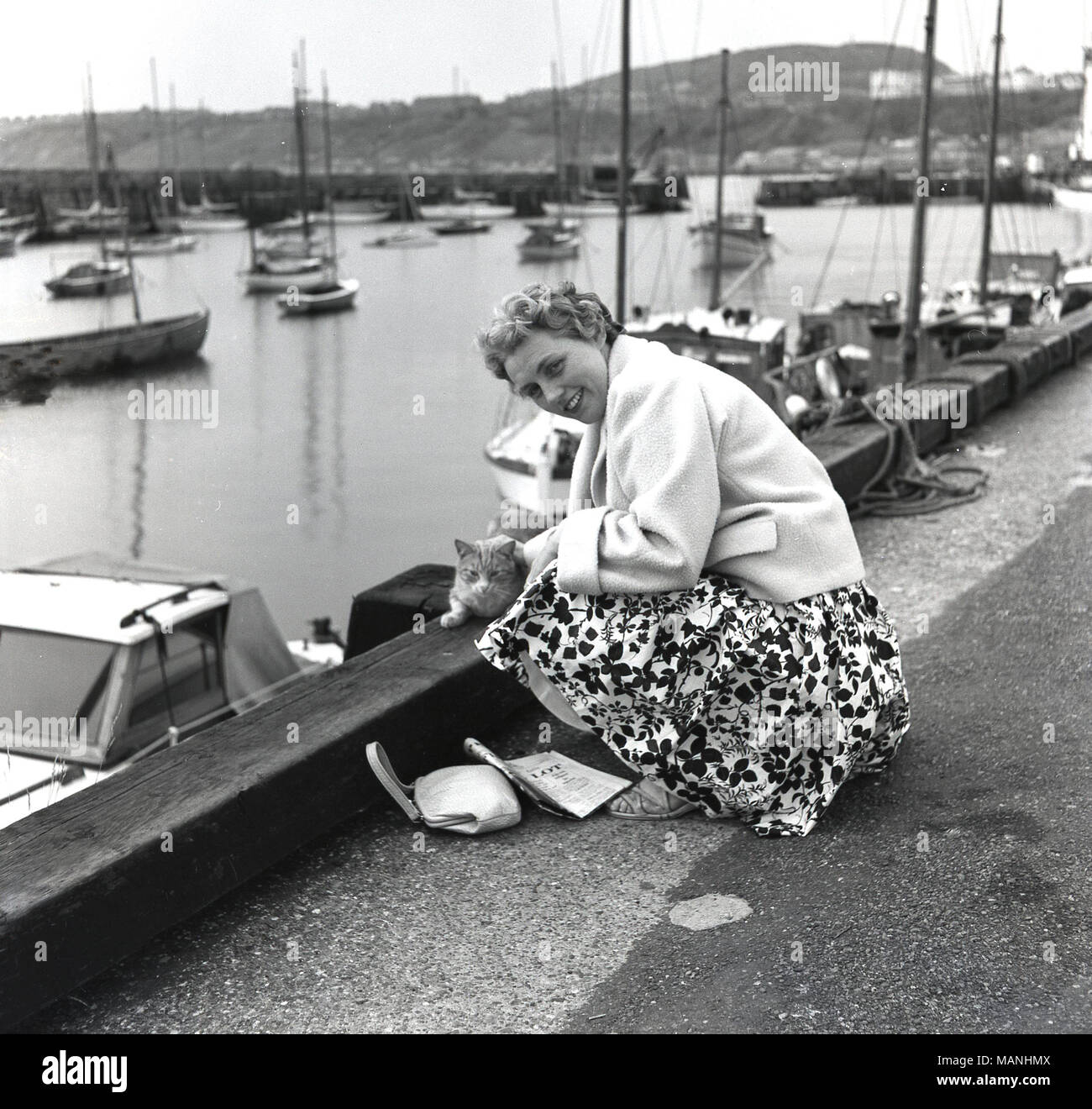 1950s, historical, a well-dressed lady on holiday kneeling by a harbour's edge stroking a local cat, England, UK Stock Photo