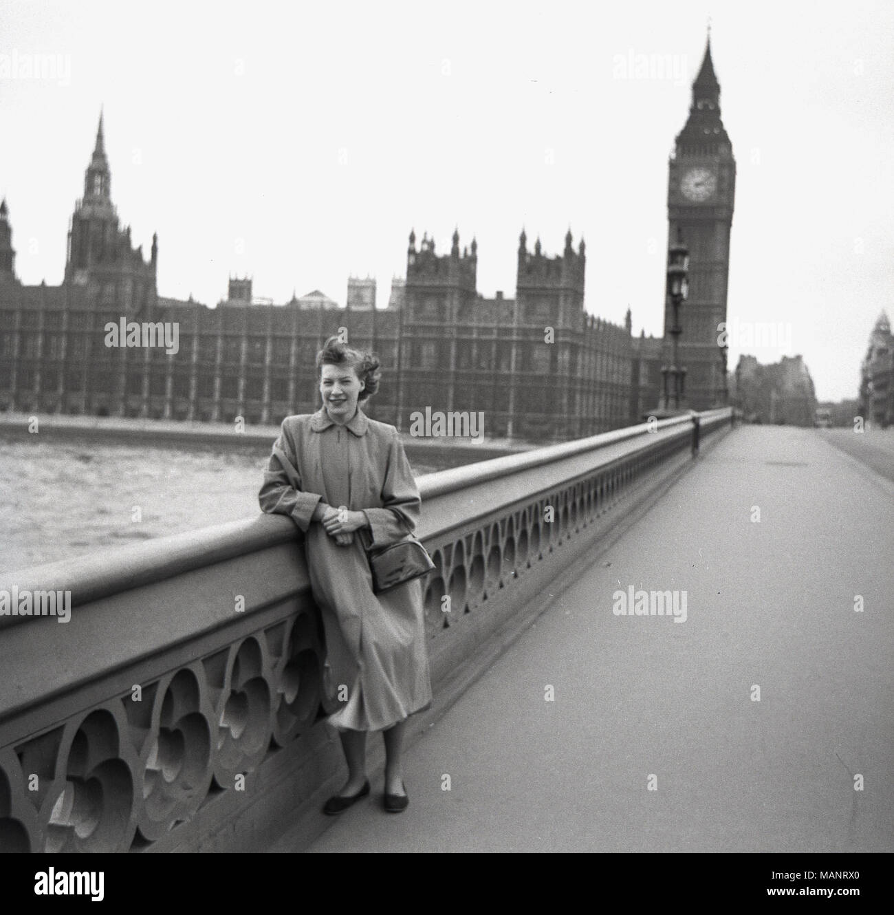 1951, historical picture of a lady standing, leaning on the safety railing or parapet on Westminster Bridge, with the UK Houses of Parliament in the background. Amazingly, given this picture was taken at daytime, the bridge is completely empty, with not a single other person anyway on this side of the bridge. Perhaps it was a Sunday morning and one can surmise that as it was not many years following WW2, and that London had very few visitors. Stock Photo