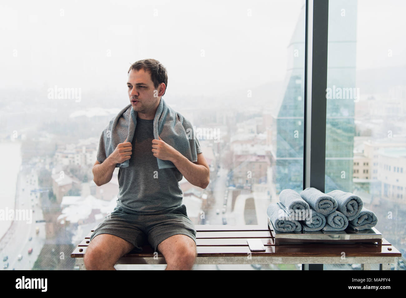 Young man in gym after workout, sitting on a bench in front of wide windows overlooking modern city skyscrapers Stock Photo