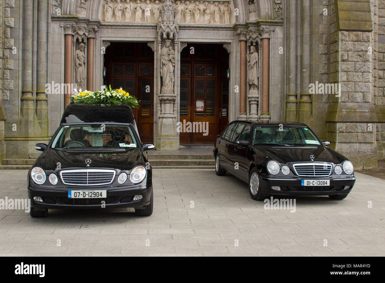 A hearse and a funeral car parked outside St Colman's Cathedral in Cobh Cork Ireland during a service for a local dignitary Stock Photo