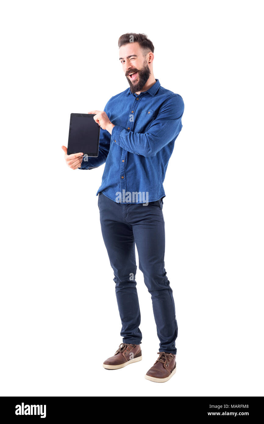 Funny expressive young bearded man presenting blank tablet screen winking at camera. Full body isolated on white background. Stock Photo