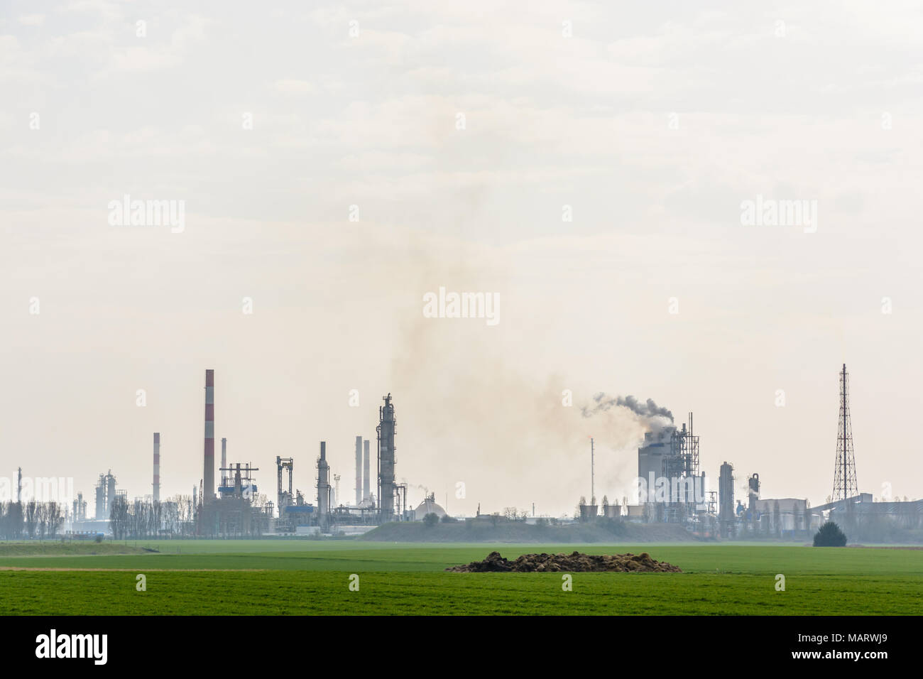 An oil refinery with smoking chimneys releasing toxic gases in the air, surrounded by crop fields in the french countryside under a pale sunlight. Stock Photo