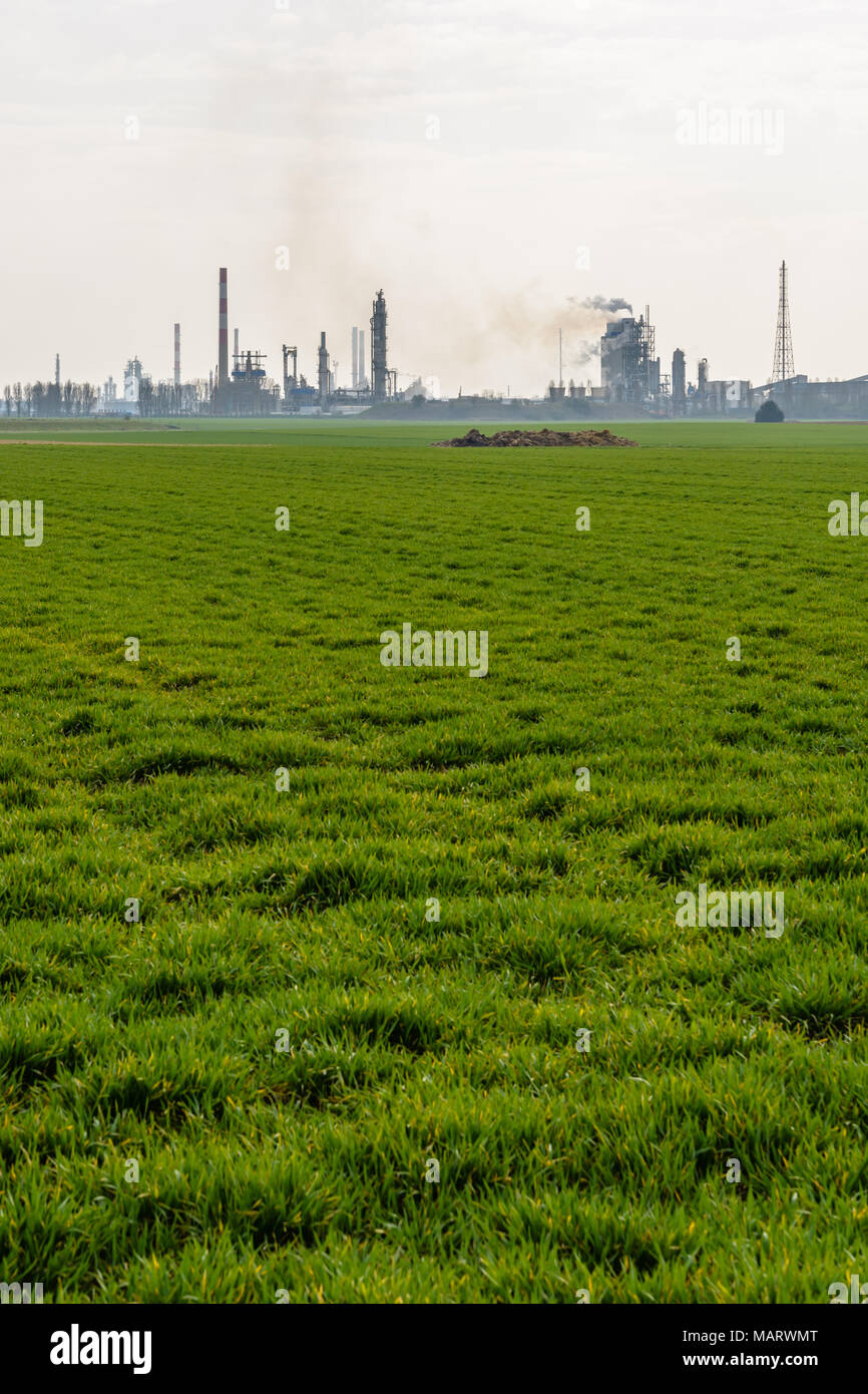 An oil refinery with smoking chimneys releasing toxic gases in the air, surrounded by crop fields in the french countryside under a pale sunlight. Stock Photo