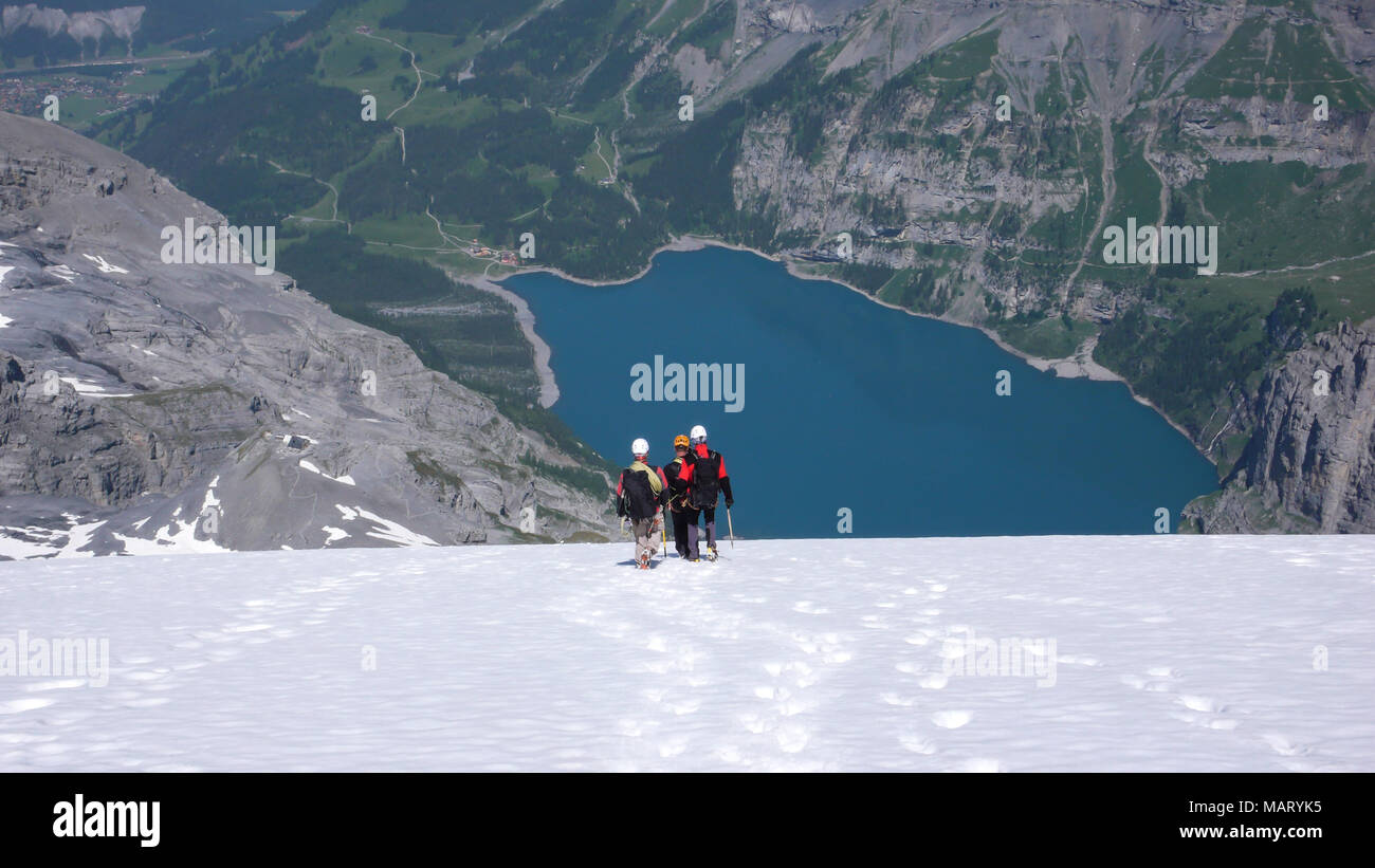 mountain guide with two clients descending a steep white glacier with a fantastic blue mountain lake far below Stock Photo